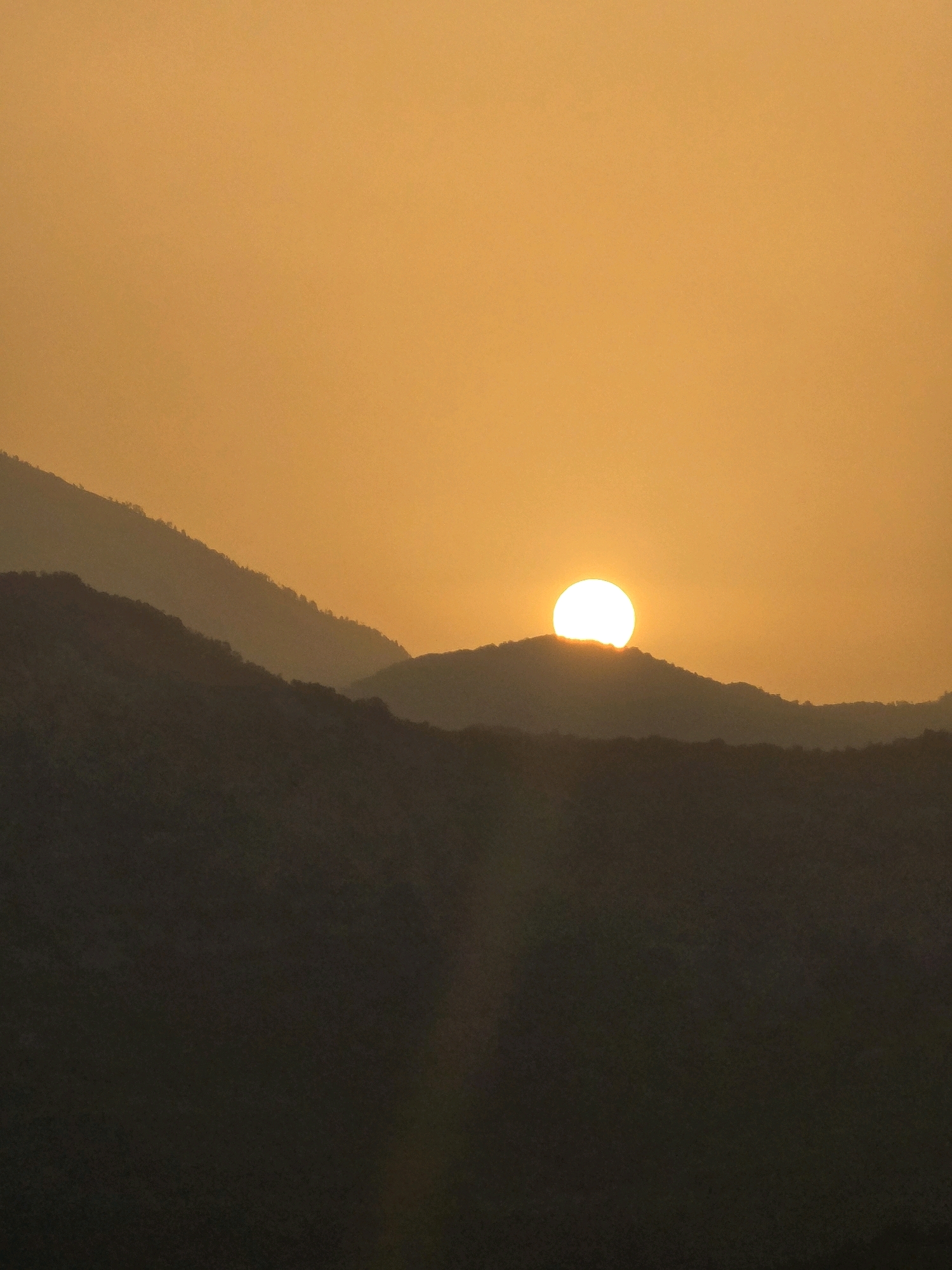 The image depicts a sunset over a mountainous landscape, likely the area surrounding Medun, Montenegro. The sun, a bright, glowing orb, is positioned just above the crest of a dark, silhouetted mountain range. The sky is a uniform, hazy orange, with the sunlight creating a soft diffusion. The foreground is dominated by layered, dark mountains that fade into the distance. There are no people or man-made structures visible in the frame, emphasizing the natural and serene environment. The overall impression is one of a peaceful, twilight scene in a rugged, natural setting.