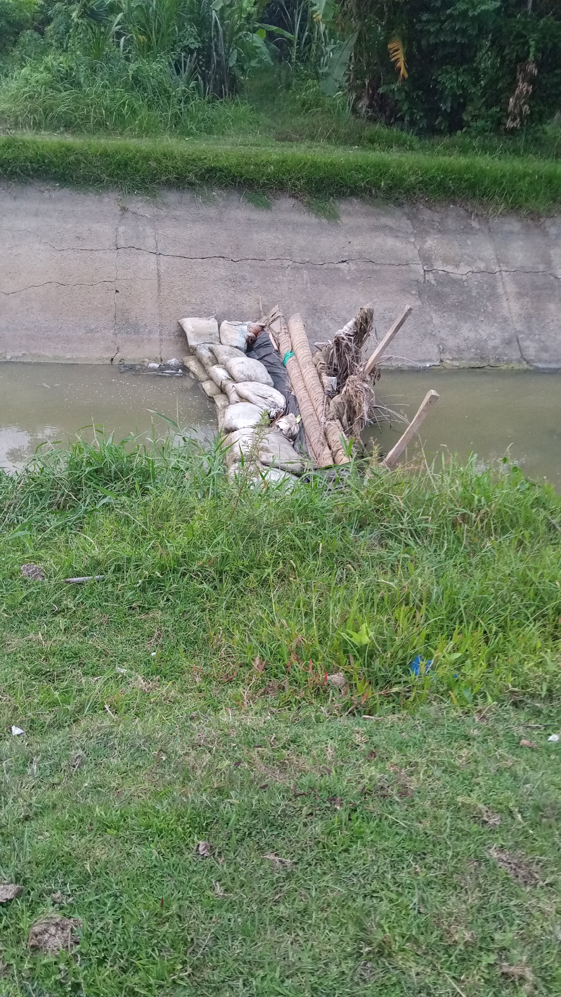 A water channel, likely an irrigation canal, is filled with brownish water. The channel's banks are made of concrete. In the middle of the channel, there is a makeshift dam or barrier constructed from sandbags, rolled woven material (possibly coconut fiber mats), pieces of wood, and other debris. The banks of the channel are covered in green grass and vegetation. The photo was taken in Uteunreutoh, Indonesia.
