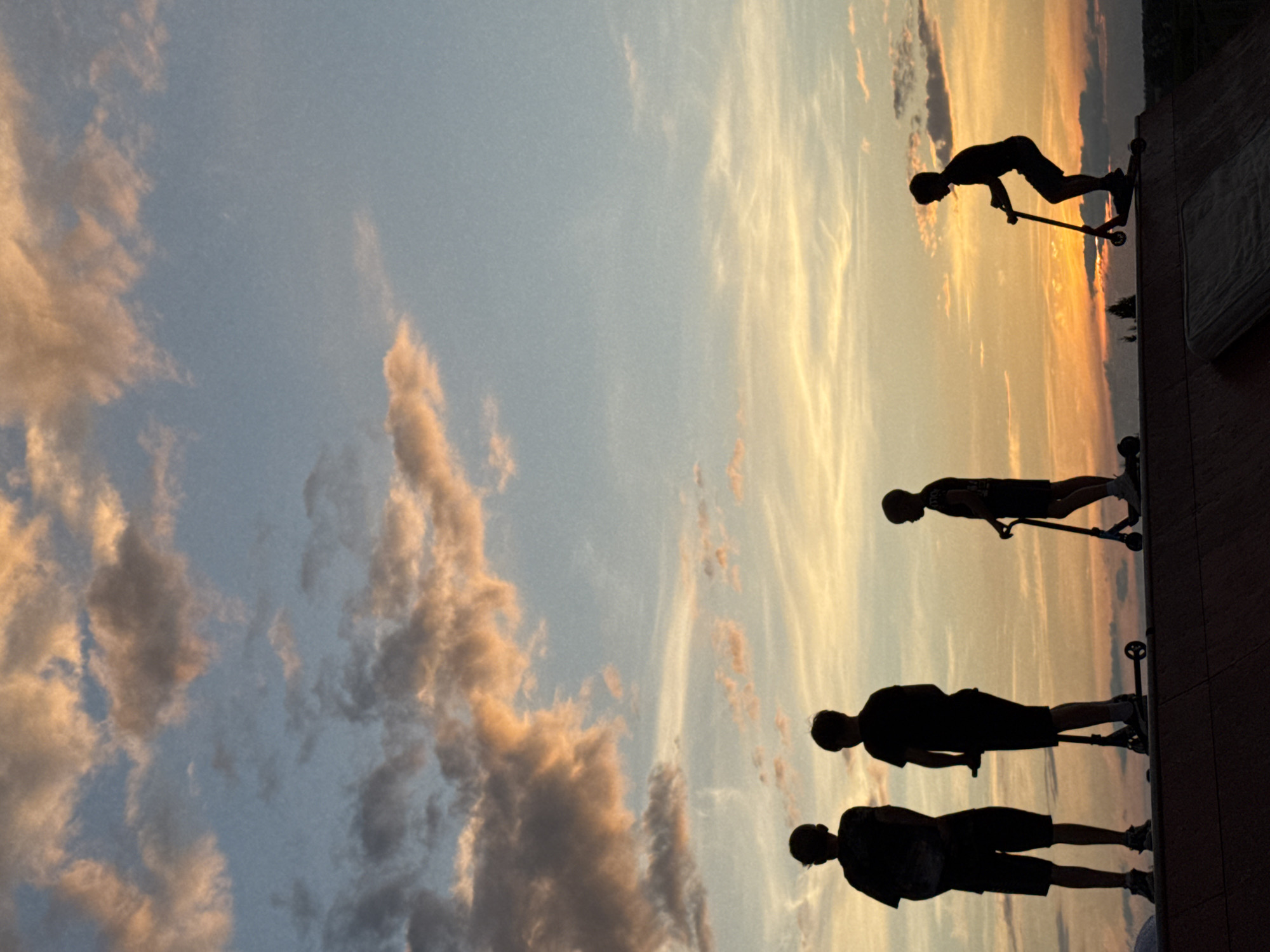 The image shows four silhouetted figures of young people on scooters at sunset. The figures are standing on a ramp or platform. The sky is filled with clouds, some of which are illuminated by the setting sun. The scene is likely a park or recreational area. The location is specified as Mikhaylovsk, Russia. The time of day is sunset, as indicated by the warm colors in the sky and the silhouettes. There are no discernible interactions between the figures. The individuals appear to be engaged in recreational activity, possibly scooter riding. The environment is outdoors, with a focus on the sky and the silhouetted figures.