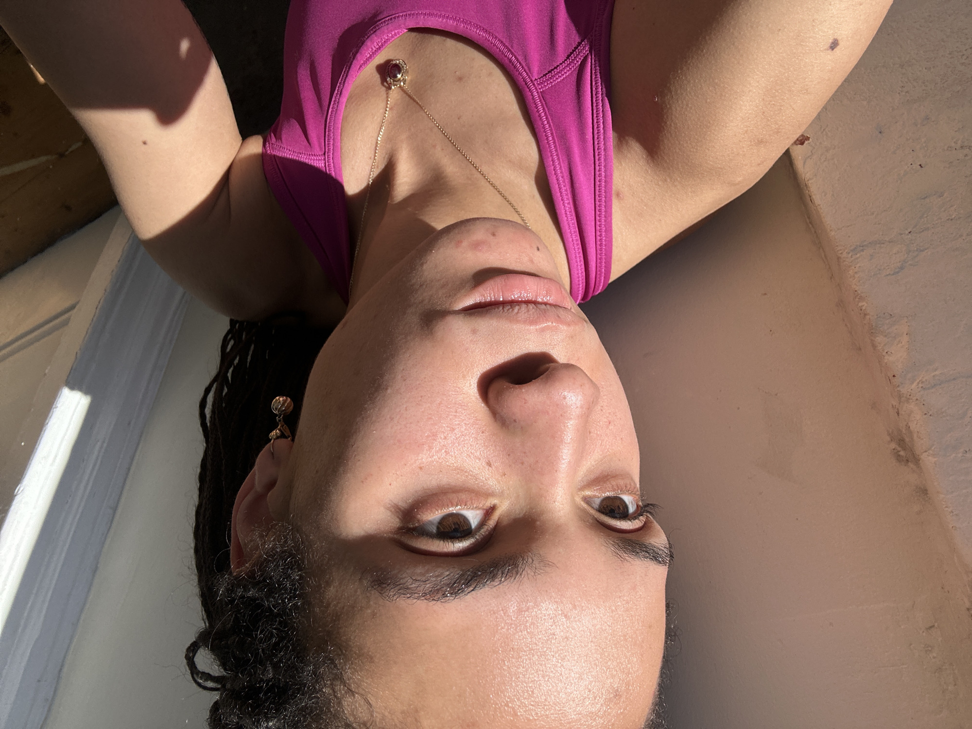 This close-up portrait features a person, likely an adult, shown upside down, implying they are lying on their back and looking directly up at the camera. The subject is wearing a vibrant magenta sports bra or tank top. A delicate gold chain necklace with a small, dark red or purple pendant rests on their chest. Dark, curly or braided hair frames the face, and a detailed gold-toned earring is visible on the left ear.
The person's face shows natural features, including dark brown eyes, full eyebrows, and slightly parted lips. Minor skin texture and blemishes are visible on the chin and forehead. The scene is illuminated by strong, warm sunlight coming from the left, casting prominent shadows across the face and upper body, indicating it is daytime, possibly morning or late afternoon.
The background is simple and somewhat abstract due to the angle, revealing a light-colored wall or ceiling on the right and a darker, possibly wooden, surface on the left. The subject's expression is neutral and direct. There is no visible text in the image. No specific location cues for London, United Kingdom, are present beyond the suggested indoor or semi-enclosed setting.