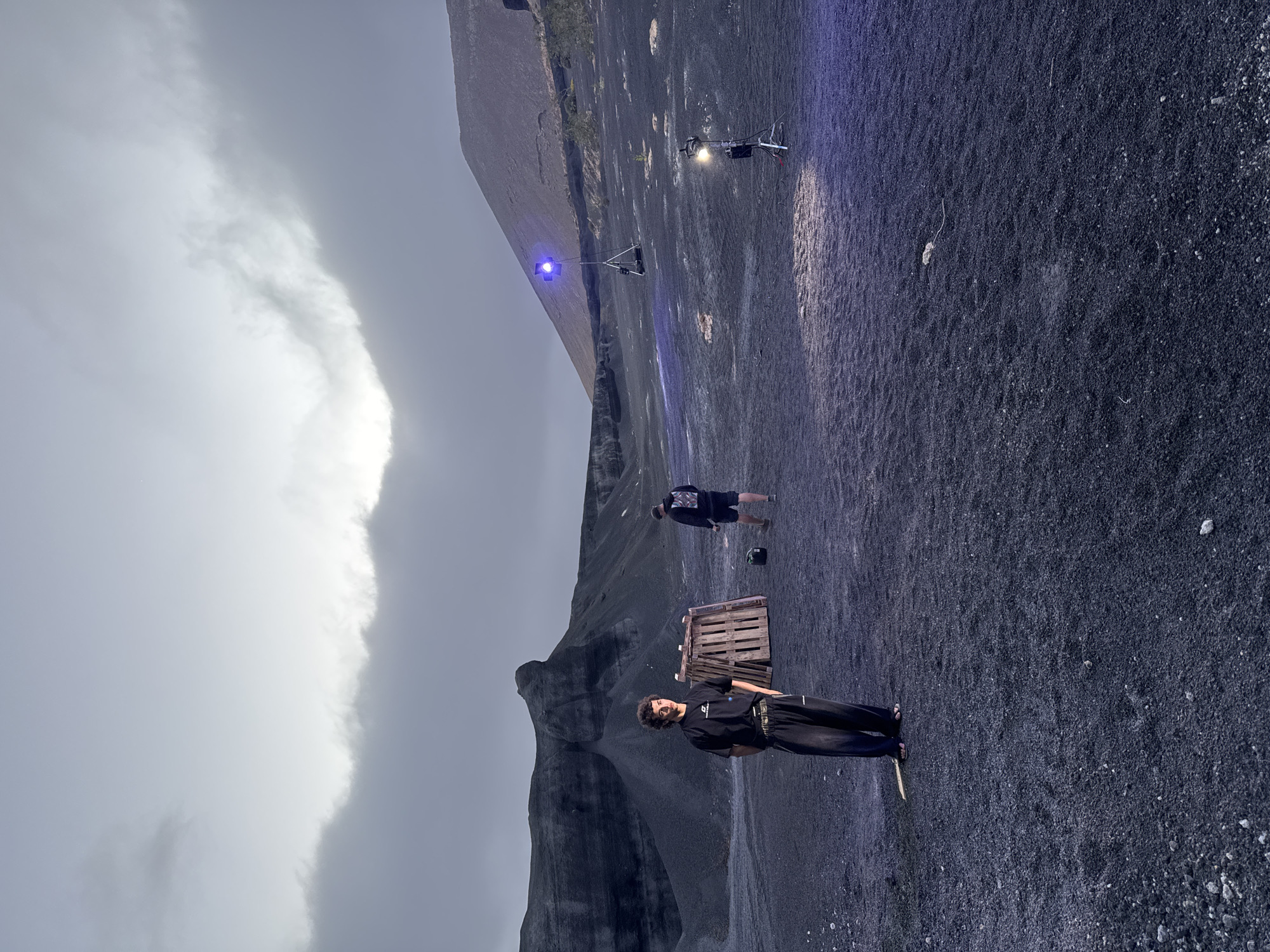 The scene is an outdoor setting with black, gravelly terrain. There is a man in the foreground wearing a black t-shirt and pants, standing near a wooden pallet. In the mid-ground, there is another person standing, appearing to be looking down, with a small object or item nearby. Further in the background are two bright lights on separate stands. One light is blue, and the other is white. Above, there are dark, cloud-filled skies. Mountains are visible in the background. It is likely the location is a landscape in Teguise, Spain.