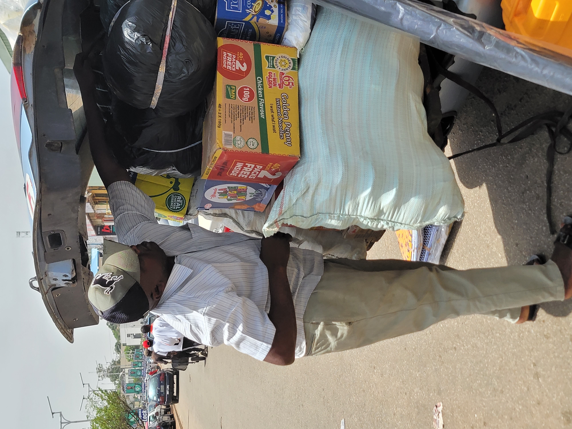An individual wearing a cap, a vertically striped shirt, and beige trousers is situated near the open hatch of a vehicle filled with various items. A box of "Golden Penny Instant Noodles" with "Chicken Flavour" is visible. Other visible objects include bags, boxes, and a yellow container. The scene is set on a paved road with other vehicles, motorcycles, and people present in the background. Signs with "Opay" and "Lock Regency Location" are also observed in the background, indicating a commercial area in Jos, Nigeria.