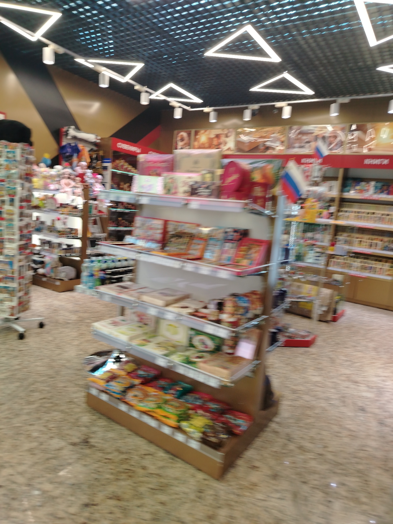 The image shows the interior of a shop, possibly a souvenir or gift shop, in Khimki, Russia. There are shelves stocked with books, postcards, toys, and other merchandise. A Russian flag is visible among the items for sale. The shop features a distinctive ceiling with triangular lighting fixtures. The floor is a speckled light brown color. The photo is taken from within the store, looking towards shelves and merchandise. The image appears slightly blurred, possibly due to motion.