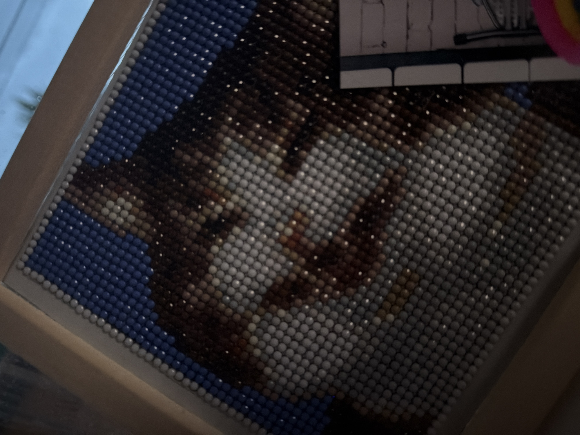 This media file presents an overhead, slightly tilted view of a finished or near-finished beaded artwork, resembling a diamond painting, set in Chaikovsky, Russia. The central subject is a pixelated depiction of an animal's face, likely a cat or a dog, rendered with thousands of small, round, spherical beads.
The animal's face is formed by an intricate arrangement of beads in various shades of brown, gray, white, and black, creating the impression of fur texture, eyes, and a nose. A significant portion of the artwork's background surrounding the animal's head is filled with dark blue beads, with a border of lighter gray or white beads along the edges of the blue section. The artwork is contained within what appears to be a light-colored wooden or plastic frame.
In the upper right corner of the image, parts of other objects are visible. There is a section of a white, rectangular item with several thin black horizontal lines, which might be a reference chart for the artwork or a stylized representation such as a piano keyboard. Adjacent to this white object, a small, vibrant circular item, possibly a crafting tool or a toy, displays colors like pink and yellow.
The overall lighting is somewhat dim, but reflections are visible on many of the beads, indicating a light source, possibly artificial or ambient window light, illuminating the piece. No specific activity is taking place within the frame, and there is no discernible text beyond the abstract lines on the white object. The image captures the intricate detail and texture of the beadwork.