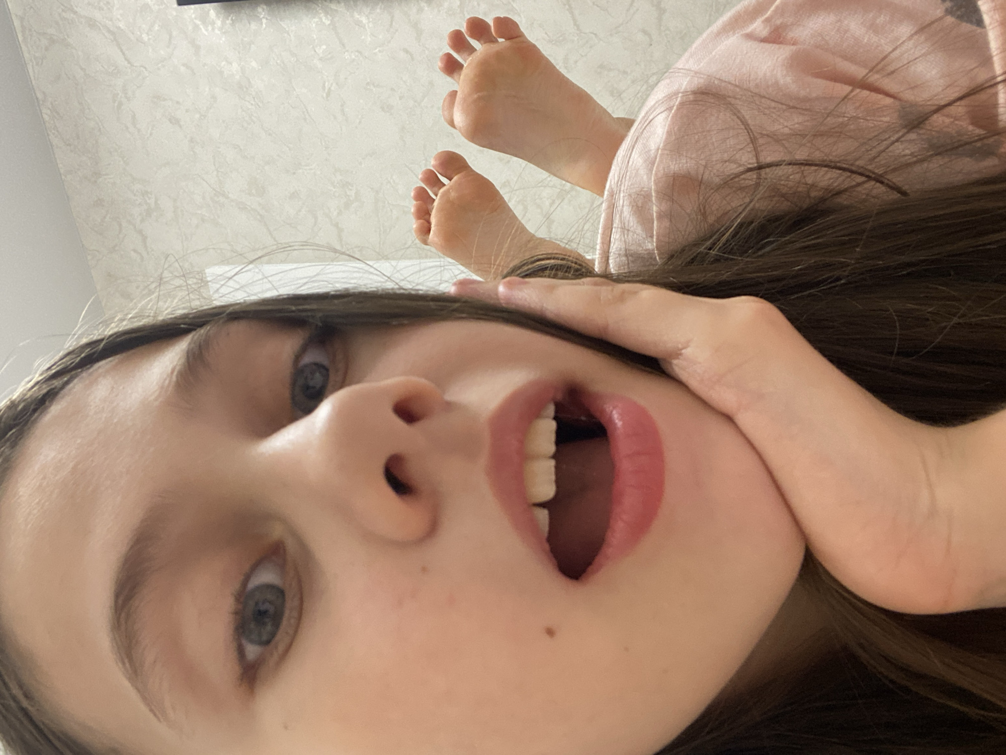 A young girl lies on her back, face looking up at the camera. Her hair is dark brown, and her eyes are gray. Her mouth is open, and her hand touches her face. Above her head and in the background, her feet are visible in the upper part of the frame. The background appears to be a ceiling with a patterned design. The location is not identifiable as Izhevsk, Russia, based on the visible details.