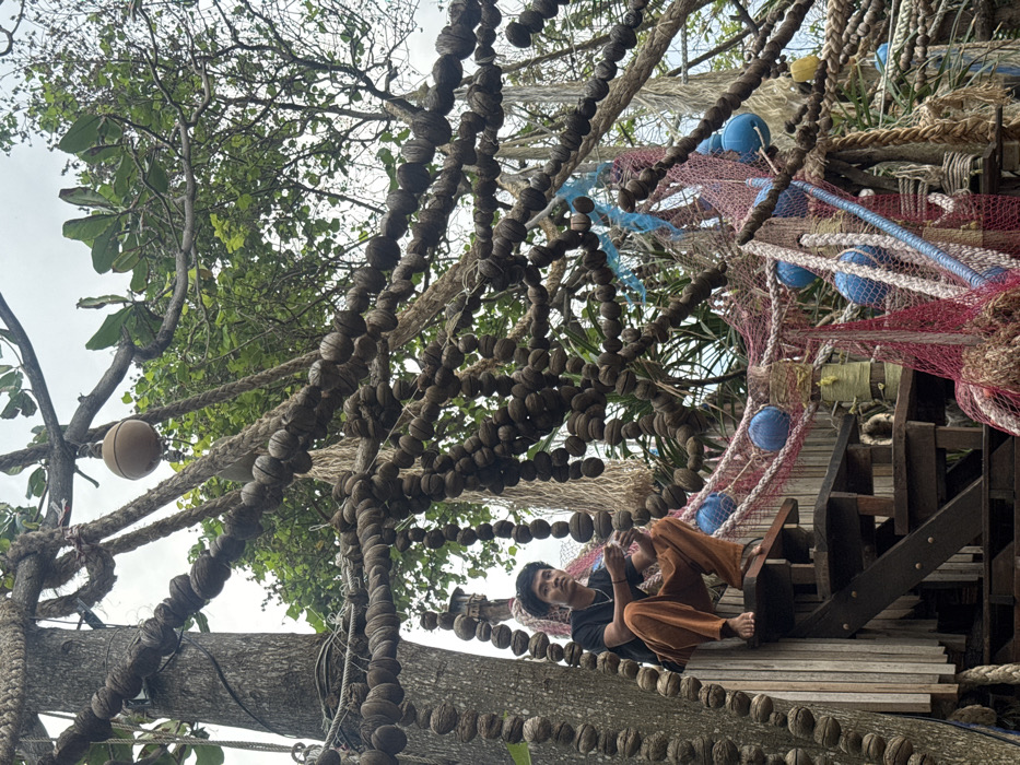 In Baan Sri Thanu, Thailand, an outdoor scene is depicted, heavily adorned with a dense array of nautical and natural elements. A single male individual, appearing to be an adult of Southeast Asian descent, is observed squatting barefoot on a wooden step of an elevated wooden structure, positioned on the left side of the frame. He wears a black short-sleeved t-shirt and loose-fitting burnt orange trousers, and maintains direct eye contact with the viewer. His left hand is slightly raised, possibly holding a small, light-colored object.
The environment is characterized by numerous thick ropes, some plain, others strung with multiple small, dark, spherical, bead-like objects, crisscrossing the upper and central areas, secured to prominent tree trunks and branches. Draped red and blue fishing nets are visible in the central and lower right, entangled with blue spherical buoys. A larger, light brown spherical buoy hangs from ropes in the upper left. Green foliage from trees forms the background, with an overcast sky visible above. The wooden structure the individual is on consists of vertical planks and a series of horizontal steps.
