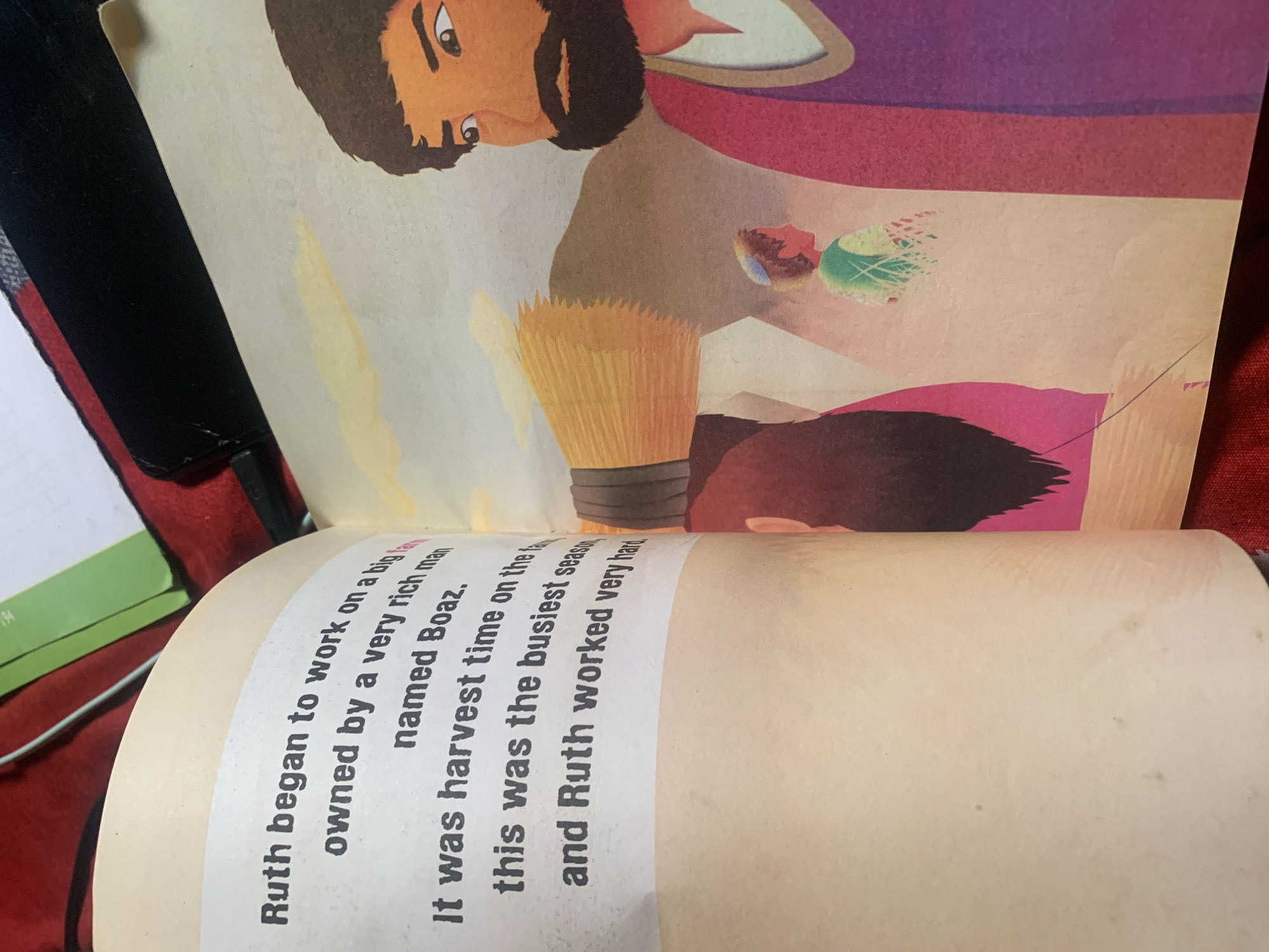 The image shows an open book with text on the left page and an illustration on the right page. The text on the left page reads: "Ruth began to work on a big farm owned by a very rich man named Boaz. It was harvest time on the farm. This was the busiest season, and Ruth worked very hard." The illustration on the right depicts a scene from the story. A man with a beard and dark hair is prominent on the right, wearing a purple robe. In the background, a younger person in a green tunic and a cap is visible. Another figure, mostly obscured, is seen to the left of the younger person, with pink and black colors suggesting clothing. The illustration has a stylized, cartoonish appearance, with warm colors and simplified forms. The overall context suggests a biblical story, likely the Book of Ruth. There is no information in the image that indicates the location is in Jauro Yinu, Nigeria.