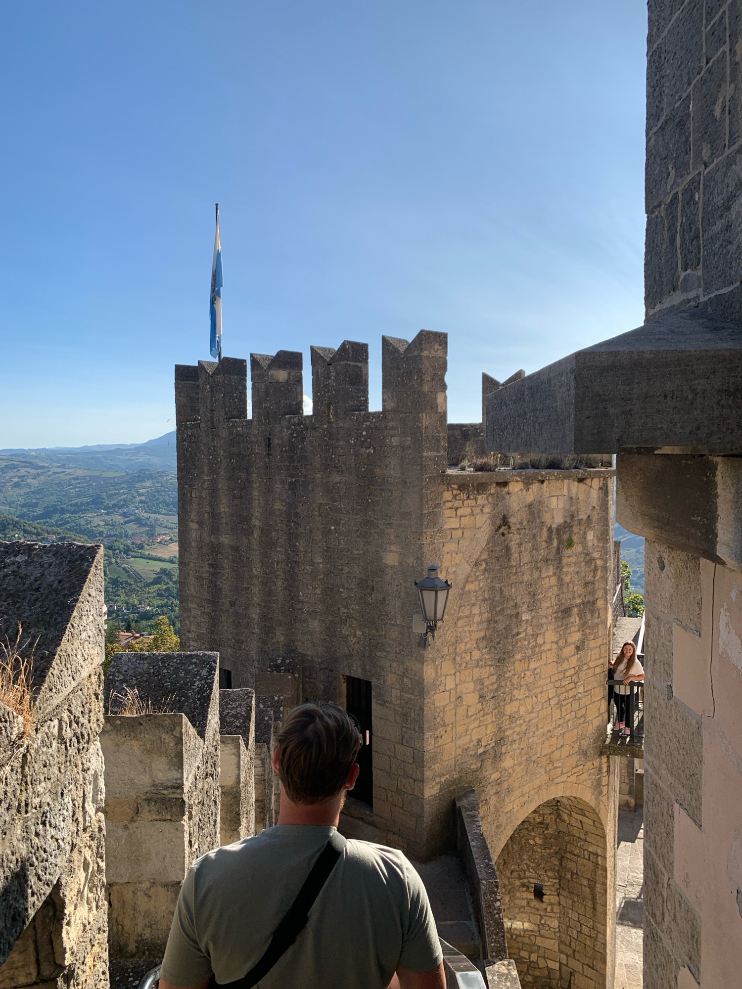 The image captures a view from a fortress in the City of San Marino, San Marino. In the foreground, the back of a man wearing a light green t-shirt and a black strap across his chest is visible as he walks down a stone staircase. The fortress walls, constructed from rough-hewn stones, dominate the scene with their crenellated battlements. A flag, featuring blue and white stripes, flies from a pole atop one of the towers. In the mid-ground, a young woman with long brown hair stands on a balcony, looking towards the camera. She is wearing a white top and dark trousers. Beyond the fortress, a verdant, rolling landscape stretches out under a clear blue sky, with distant hills and scattered buildings. The architecture suggests an ancient, historical location, typical of San Marino's fortifications.