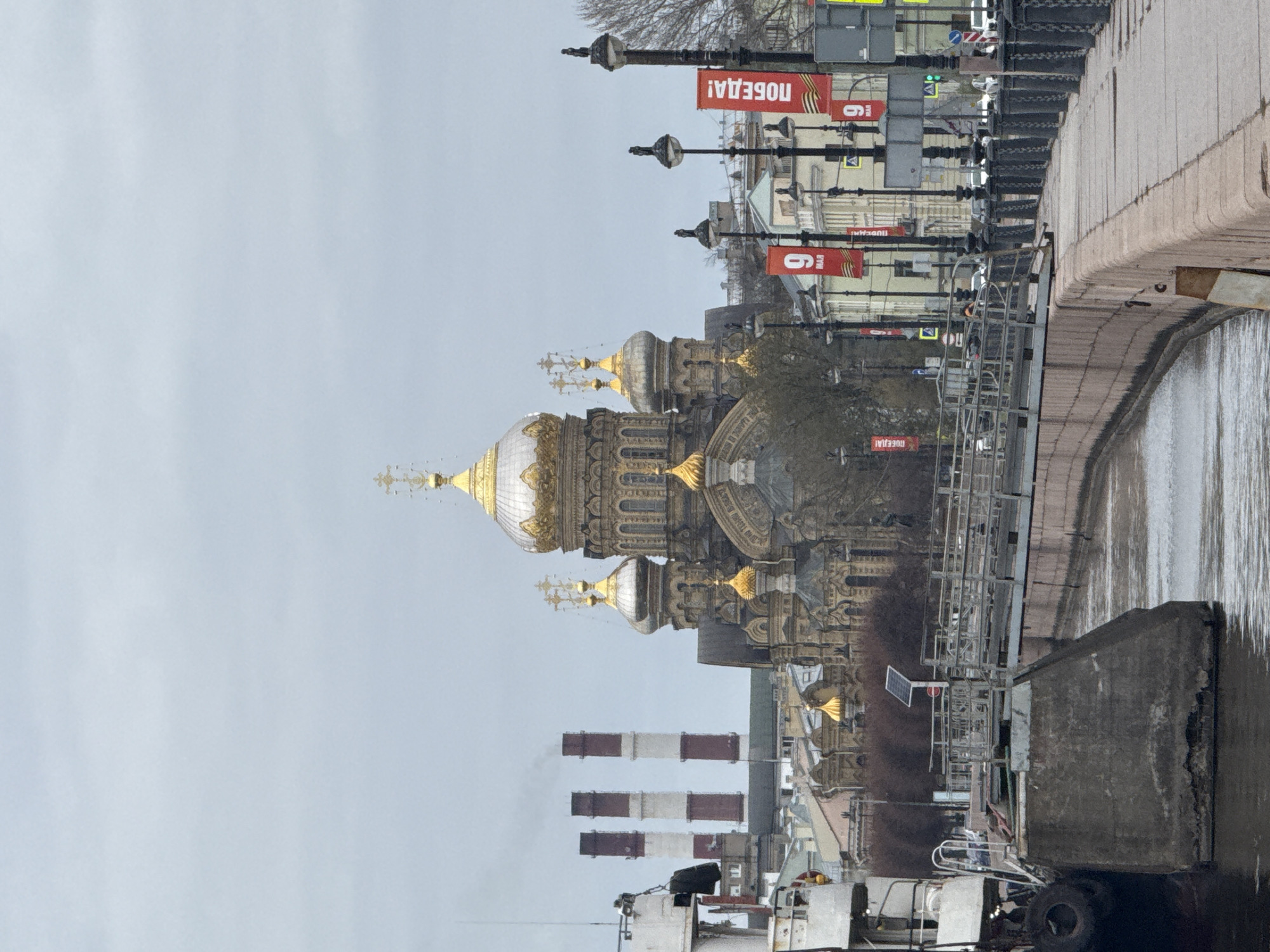 The image presents an urban landscape in Saint Petersburg, Russia, featuring a grand, ornate cathedral as a central subject. This cathedral is distinguished by its multiple golden, bulbous domes and intricate architectural details on its facade.
To the right of the cathedral, a canal or river runs, bordered by a substantial concrete embankment. A bridge with dark metal railings spans the waterway further back. In the foreground on the canal, a section of a concrete dock or barge is visible, along with parts of a work boat or industrial vessel, including large tires and metal frameworks. Three tall, striped industrial smokestacks—alternating red and white—rise prominently in the foreground, emitting faint plumes of smoke.
To the left of the cathedral, a street is visible, lined with streetlights from which hang red banners. These banners display white text, clearly reading "ПОБЕДА!" (Pobeda!), which translates to "VICTORY!", alongside the number "9", signifying May 9th (Victory Day). Bare trees are scattered throughout the scene, suggesting a colder season.
The sky is overcast and uniformly light grey, indicating a daytime setting without direct sunlight. The overall atmosphere is quiet and somewhat muted due to the weather and lack of visible human activity. The scene offers a view blending historical architecture, modern urban infrastructure, and industrial elements within the city context.