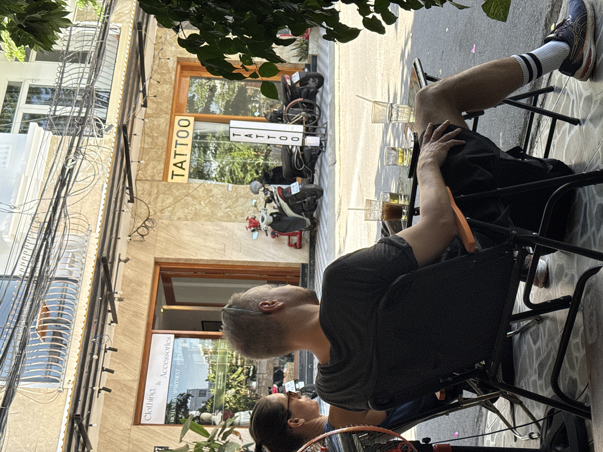 Two individuals, a woman with dark hair pulled back and sunglasses, and a man with short blond hair and sunglasses perched on his head, are seated at a table outside a shop. The man is wearing a dark t-shirt and shorts, and the woman is wearing a dark tank top. They are both looking away from the camera. On the table in front of them are three glasses with drinks and straws.
The shop behind them has two storefronts. The left storefront has a sign that reads "Clothing & Accessories" and "RIO LUXURY APARTMENT". The right storefront has a sign that reads "TATTOO" in large yellow letters. Both storefronts have large glass windows and wooden doors.
In front of the tattoo shop, several motorcycles are parked. A white sign with the word "TATTOO" written vertically in white letters on a green background stands on the sidewalk.
The environment appears to be an urban street scene in Đà Nẵng, Vietnam, with power lines strung across the top of the buildings. The buildings are made of light-colored stone, and there is greenery visible in the background and foreground. The ground is tiled with a grey pattern. The lighting suggests it is daytime.