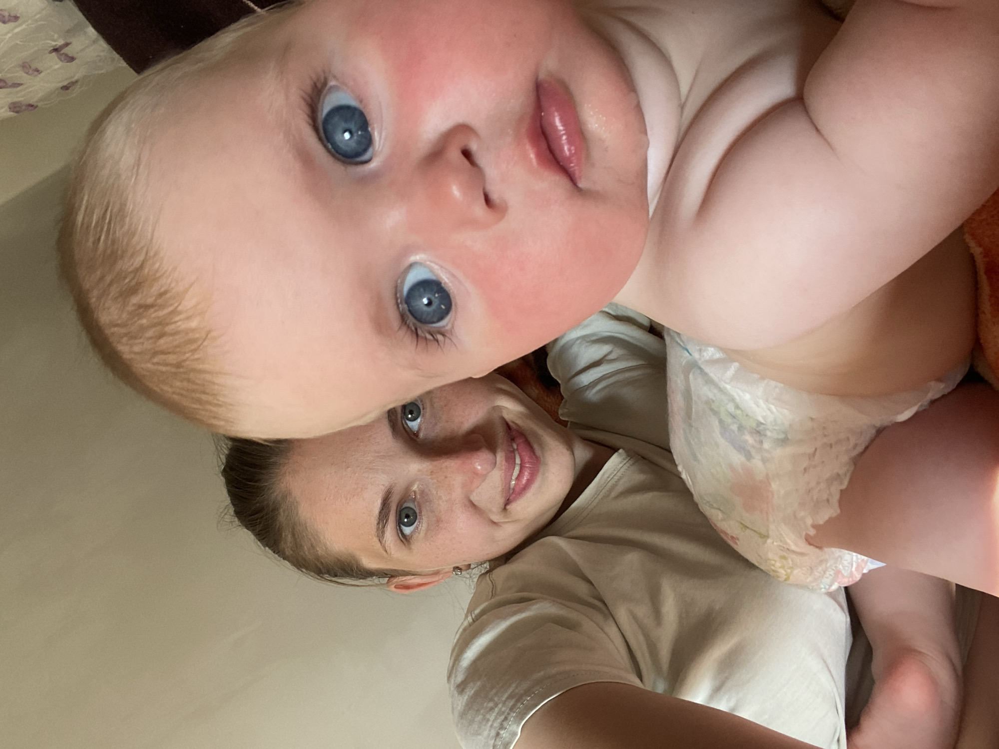 This is a close-up, vertically oriented photograph of a mother and her baby. The baby's face is prominent in the foreground, looking directly at the camera with bright blue eyes and pink cheeks. The baby's mouth is slightly open, revealing plump lips. The baby is wearing a diaper with a floral pattern. The mother is in the background, smiling and looking towards the camera. Her eyes are also blue, and she has a scattering of freckles across her nose and cheeks. She is wearing a plain, light-colored t-shirt. The background is blurred, but appears to be an indoor setting with soft, neutral-toned surfaces. The lighting suggests it is daytime, possibly indoors with natural light. The overall mood is warm and affectionate, capturing a moment of connection between mother and child.