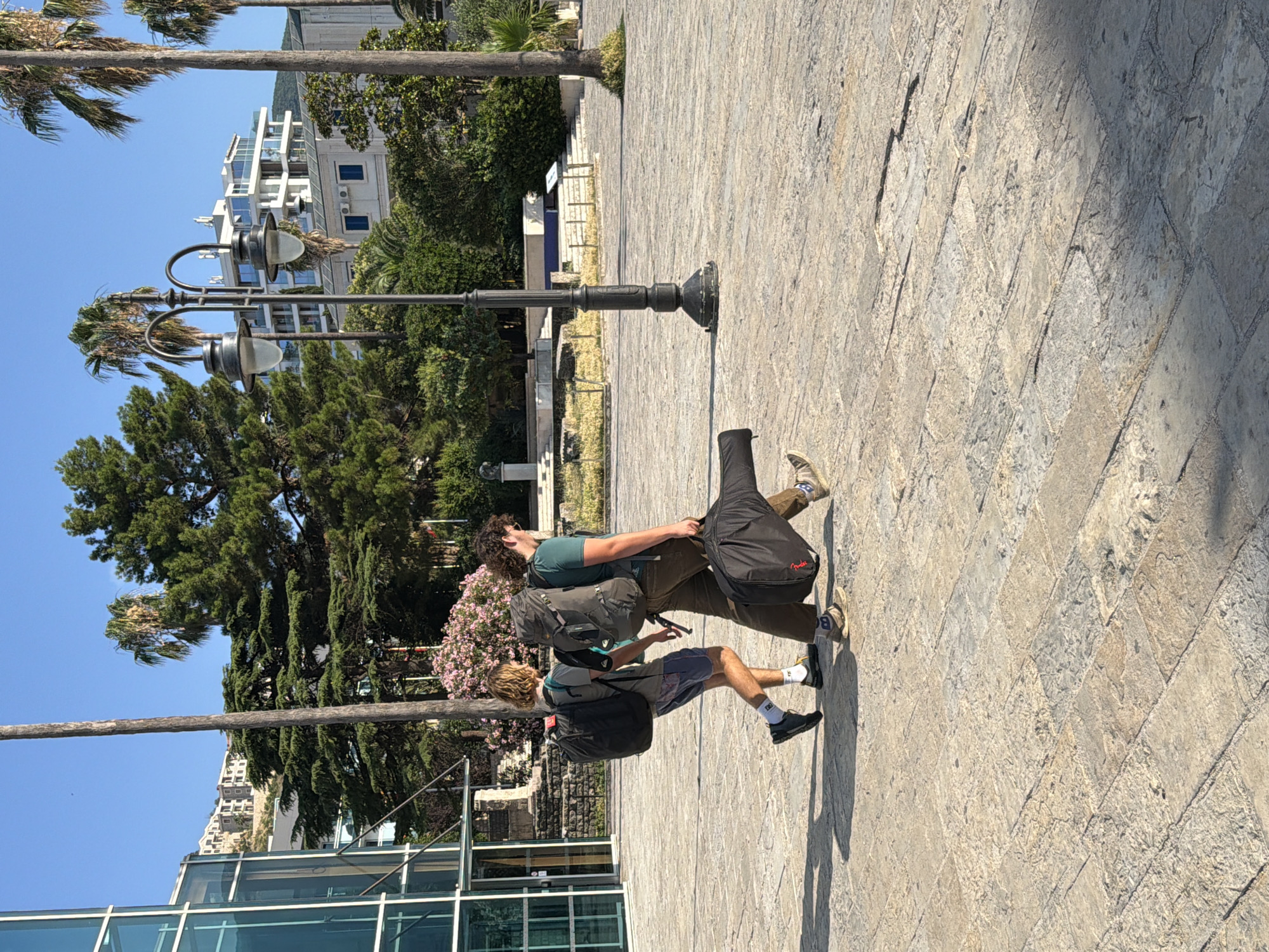 Two young men walk across a sun-drenched stone plaza in Budva, Montenegro. The man on the left wears a black backpack, blue shorts, and dark sneakers. He has light brown, curly hair. The man on the right carries a large, dark instrument case in his right hand and wears a green t-shirt, brown pants, and light-colored sneakers. He has dark curly hair. Behind them, a tall, dark streetlamp stands to the right. The plaza is paved with textured, light-colored stone tiles. In the background, a modern building with glass facades is visible on the left, and further back, a white building with balconies sits on a hillside, surrounded by greenery and palm trees. The sky is a clear, bright blue.