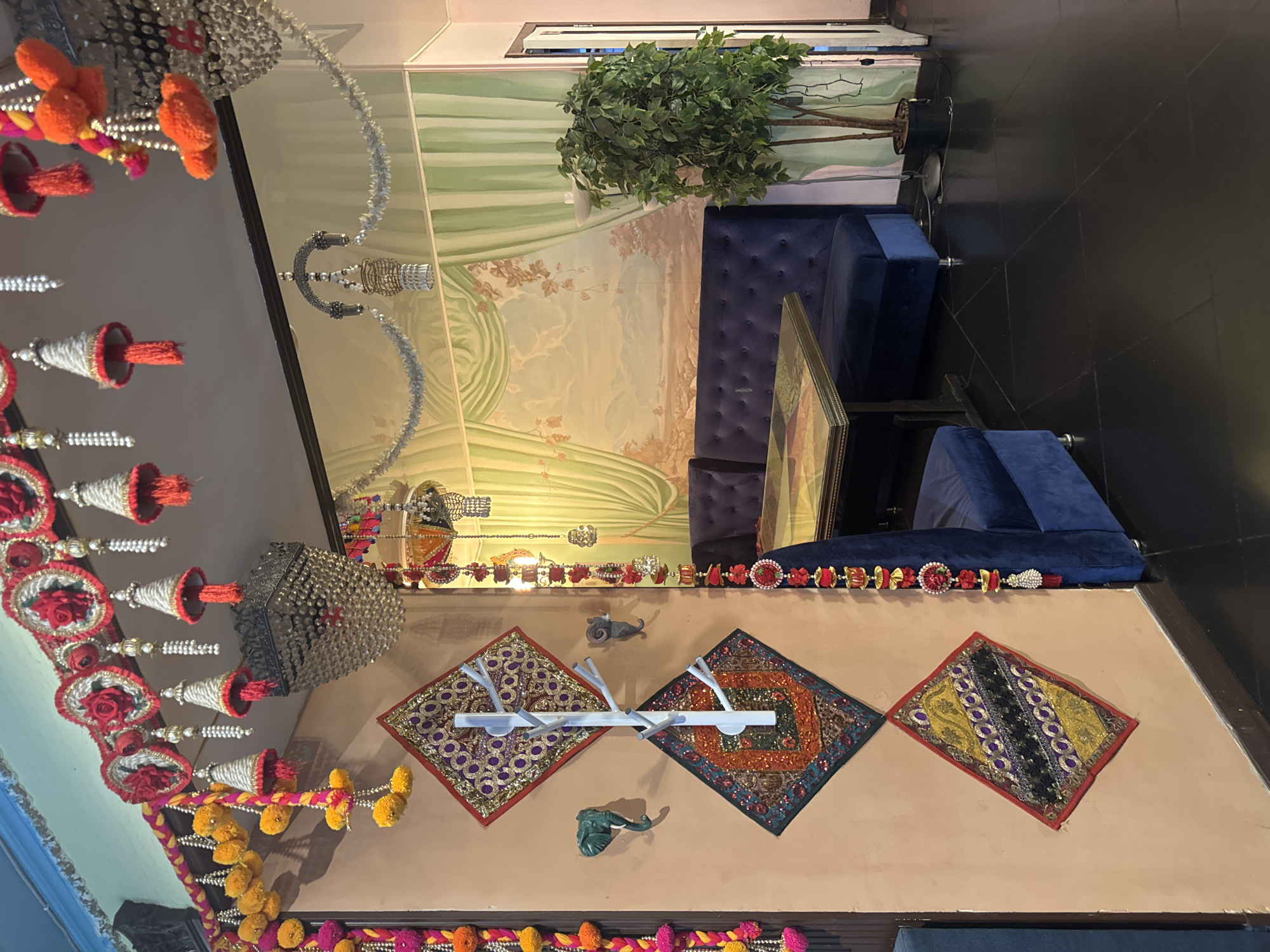 The image depicts the interior of a restaurant in Saint Petersburg, Russia, styled with a vibrant and festive Indian theme. The foreground shows a wall adorned with colorful textile decorations, including beaded garlands, pom-poms, and embroidered diamond-shaped hangings. A white coat rack with branching arms is mounted on the wall, with two small decorative animal heads mounted to its sides. Hanging from the ceiling and wall are various ornate chandeliers and decorative tassels, adding to the festive atmosphere.
The middle ground features a seating area with deep blue velvet banquettes surrounding a table with a decorative glass top. A large artificial plant stands in a black pot near the corner of the seating area. Behind the seating area, a mural on the wall depicts a pastoral landscape with rolling hills, trees, and draped curtains. The floor is made of dark, polished tiles arranged in a geometric pattern.
In the background, a doorway is visible, leading to another part of the establishment. The overall impression is of a lively and elaborately decorated space, likely intended to create an immersive cultural experience for diners.