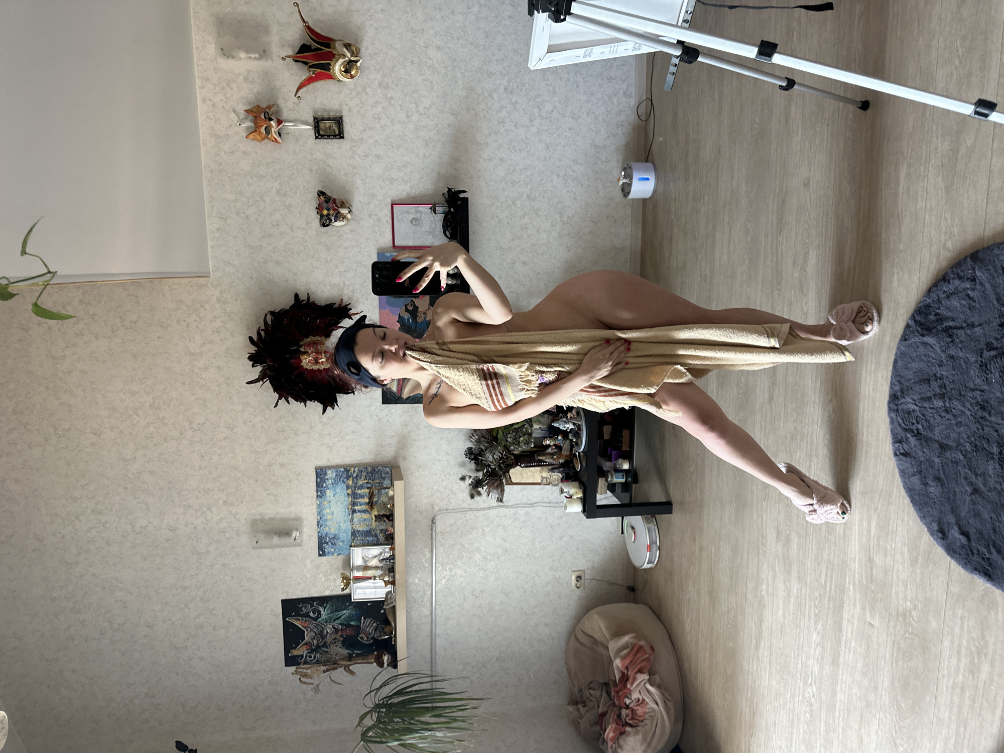 The vertical, full-shot indoor image shows a woman with dark hair and a feathered headband, appearing to be taking a selfie. She is partially covered by a light-colored towel or fabric, revealing her bare back and legs. Her left hand, with painted nails, holds a smartphone, while her right hand is holding the towel to her mouth. She is wearing light pink slippers on her feet.
The setting appears to be a room, possibly a bedroom or living area. The walls are light-colored and textured, adorned with several decorative items, including three carnival-style masks and two framed pictures or artworks. On the left side of the image, a green plant is visible.
To the right, there is a tall, light-colored stand or tripod with a white panel attached to the top, and a white, cylindrical humidifier with a blue light on its top is on the floor nearby. In the background, there's a dark-colored shelf unit holding various small objects. Another two artworks or pictures are mounted on the wall below the shelf. The flooring is light-colored wood. A dark grey circular rug is visible on the floor on the right side of the image. The time of day is not discernible, but the indoor lighting suggests it is not heavily reliant on natural light. No visible text is present in the image.