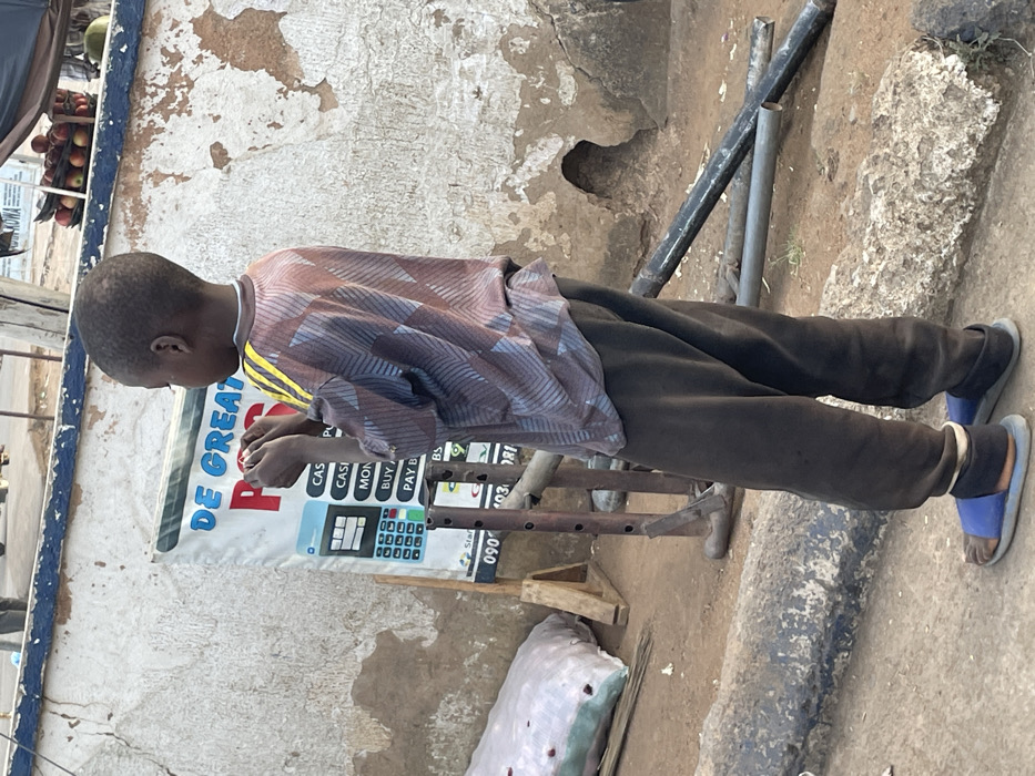 A young boy stands in front of a weathered wall next to a sign that reads "DE GREAT P" with information about cash, payment options, and phone numbers. He is wearing a patterned shirt, dark pants, and blue sandals. A rack-like structure and metal pipes are near him. To the left, a display of fruits under a cover is partially visible. The surrounding environment appears to be a street or a sidewalk.