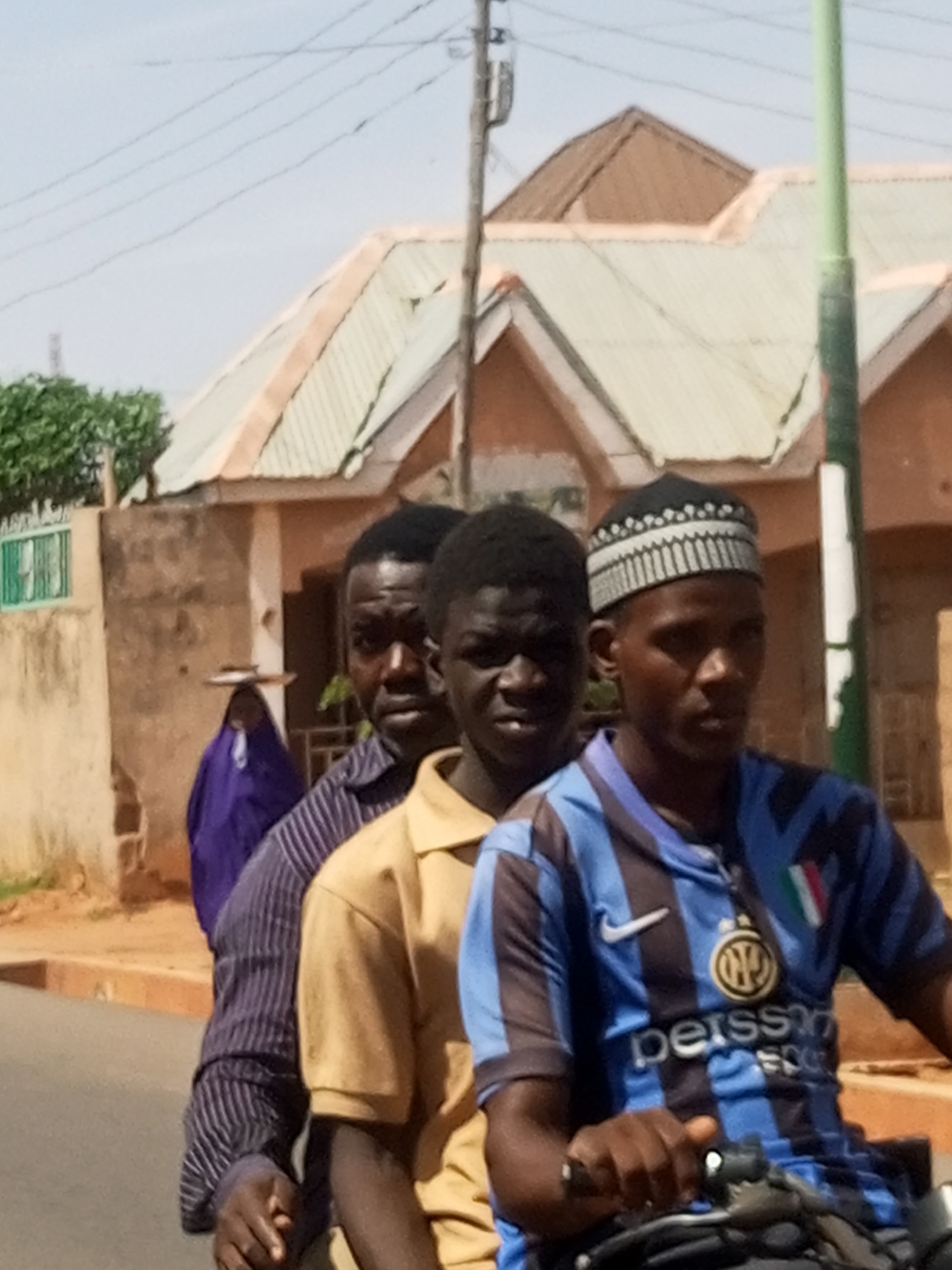 This image captures a street scene in Tempure, Nigeria, featuring three men on a motorcycle and a woman in the background. The men are positioned in a line, suggesting they are riding together, possibly on a single motorcycle, though only the handlebars of the front rider's vehicle are visible. The man in front wears a blue and black striped football jersey with a white Nike swoosh, an Inter Milan club logo, and the word "Pirelli" (partially obscured) and "Persson" printed on it, along with a small Italian flag emblem. He also wears a black and white patterned kufi cap. The second man wears a tan or light brown short-sleeved collared shirt. The third man, mostly obscured by the second, wears a dark striped long-sleeved shirt. All three men have dark complexions and short, dark hair.
In the background, a woman wearing a purple garment and a broad-brimmed hat is visible, carrying an object (possibly a tray or bowl) on her head. The setting includes several single-story buildings with corrugated metal roofs, indicative of residential structures. Power lines crisscross the upper part of the frame, and a green and white striped pole stands prominently on the right. The sky is bright and clear, suggesting daytime. The overall lighting is strong, creating some shadows. The men's expressions are neutral, focusing forward. The visible text on the jersey confirms it's an Inter Milan football club kit.
