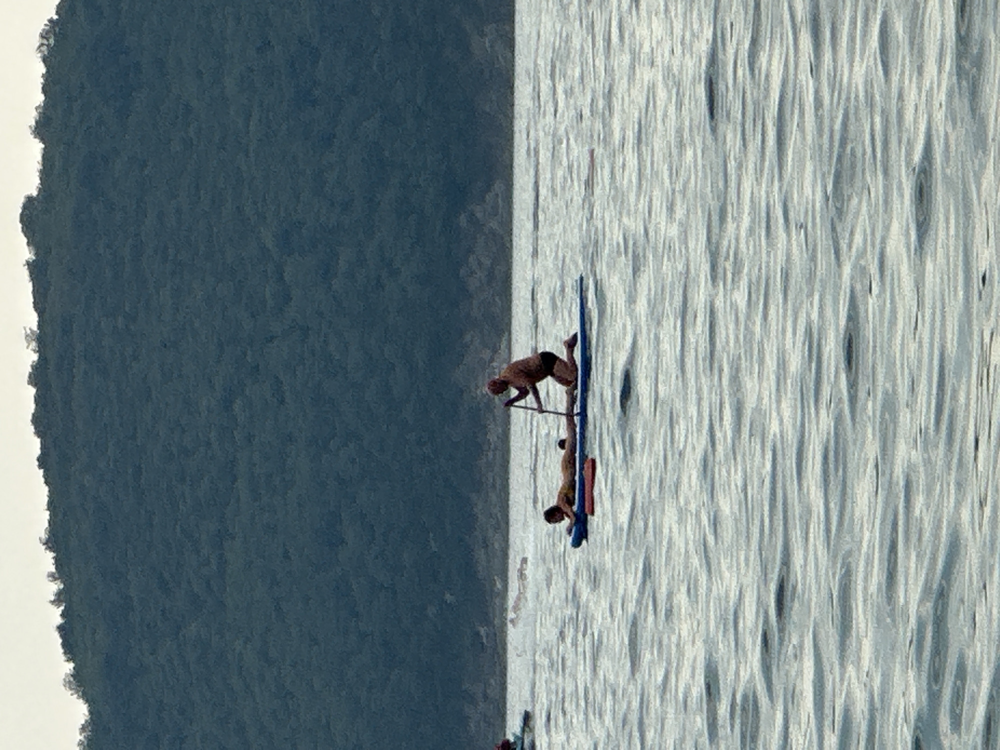 Two individuals are paddleboarding on the water near a tree-covered hill. One person is kneeling on the paddleboard, holding a paddle. The other person is lying on their stomach on the paddleboard. The water has ripples, and the sky is overcast. The scene is located near the city of Boreti, Montenegro.