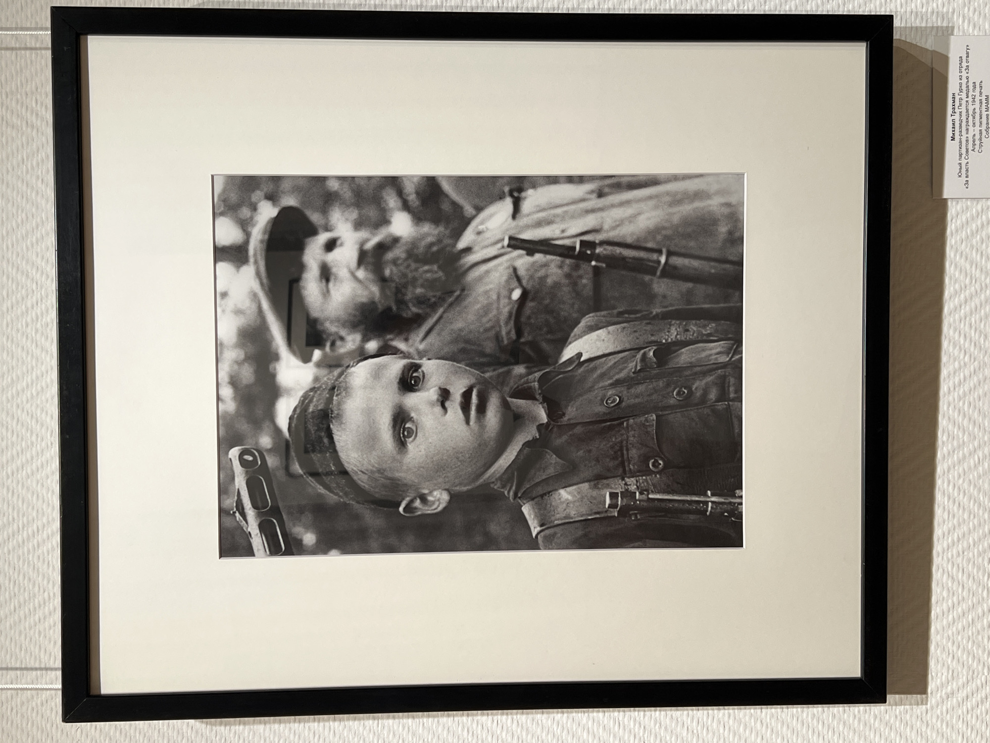The image is a black and white photograph framed in a black border and mounted on a textured off-white wall. The photograph depicts two individuals: a young boy in the foreground and an older man in the background. The boy, facing the camera, is wearing a dark shirt and a cap. He has a stern expression and appears to be carrying some sort of gear or equipment over his shoulder, possibly a rifle strap. The older man, to the right and slightly behind the boy, has a beard and is wearing a cap and a jacket. He also appears to be holding a rifle. Both individuals seem to be outdoors, with a blurred natural background suggesting trees or foliage. Below the photograph, a small white label with black text provides information about the image. The text indicates the photographer's name, "Михаил Трахман" (Mikhail Trakhman), and describes the subject as "Юный партизан-разведчик Петр Гурко из отряда «За власть Советов», награждается медалью «За отвагу»" (Young partisan-scout Petr Gurko from the detachment "For the Power of Soviets," awarded the medal "For Courage"). It also states the date as "Апрель - сентябрь 1942 года" (April - September 1942) and mentions "Струпная пигментная печать" (Pigment print). The collection is identified as "Собрание МАММ" (Collection of MAMU). The wall texture appears to be consistent with an exhibition space. There is no visible indication of the city of Novosibirsk in the image itself or the provided label.