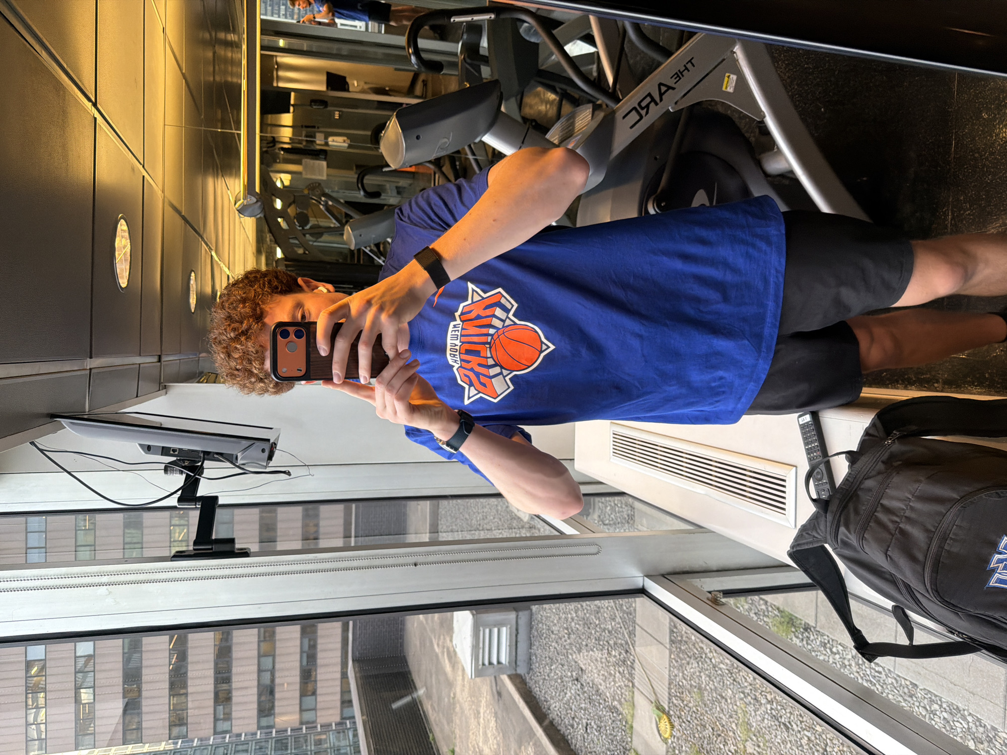 A person is standing in what appears to be a gym, possibly in the city of New York, United States of America, evidenced by the New York Knicks shirt they are wearing and the urban landscape visible through the window. The individual is holding a smartphone, presumably taking a selfie or photo, with their face partially obscured by the phone. They are wearing a blue New York Knicks t-shirt, black shorts, and a black smartwatch or fitness tracker on their left wrist.
Behind the person is a treadmill, which indicates the setting is a fitness center. Adjacent to the person, along the window, is a black backpack, a remote control, and a white radiator-like structure. A monitor is mounted near the window, displaying cabling and hardware.
The outside environment visible through the window includes buildings that suggest a dense urban area. The overall scene indicates the person is in an indoor location, possibly a gym within a building overlooking a city landscape.