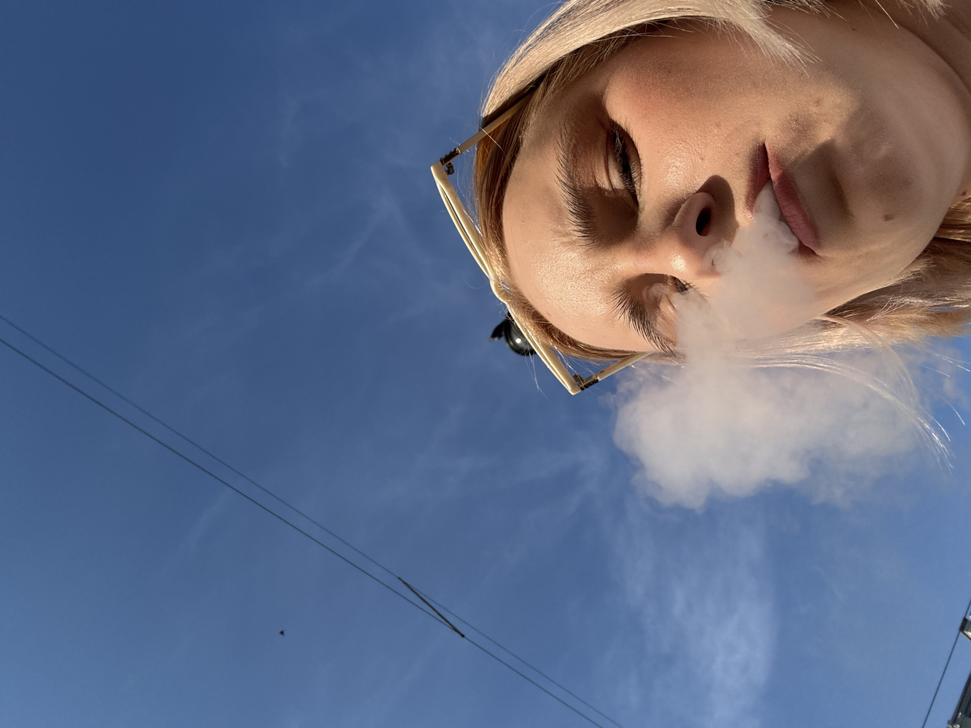 An outdoor, low-angle photograph captures a young Caucasian woman exhaling a dense cloud of white smoke against a bright blue sky, in what is identified as Saint Petersburg, Russia. The woman's face occupies the lower right portion of the frame, illuminated by direct sunlight, casting subtle shadows. She has light-colored hair and prominent eyebrows; her eyes are partially closed or squinting. Square-framed, light-colored sunglasses are perched on top of her head. The upper and left sections of the image feature a clear blue sky with some faint cirrus clouds. Two parallel overhead utility wires diagonally traverse the upper left quadrant of the sky. A small, dark, indistinct object, likely a bird, is visible in the sky below the wires in the upper left. A subtle, dark outline of a structure or building is barely discernible along the extreme bottom edge of the frame.