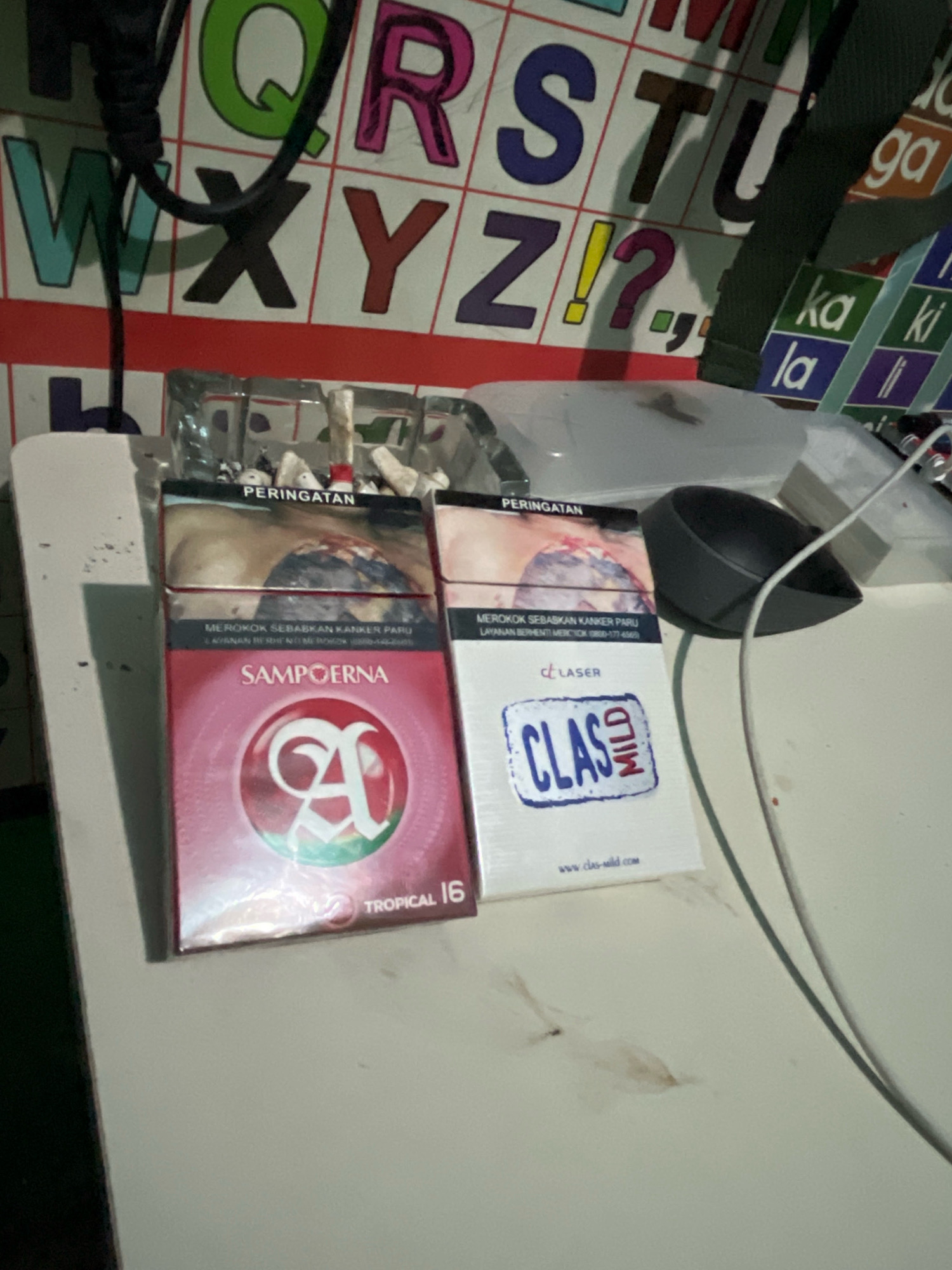 In Talang Kelapa, Indonesia, a photograph captures a table with two packs of cigarettes and an ashtray. The left pack is red and features the brand "SAMPOERNA" with a logo that appears to be an "A". It is labeled "TROPICAL 16". The right pack is white and displays the brand "CLASS MILD" with the word "LASER" above it. The ashtray, made of clear glass, contains cigarette butts. A black power cord is visible in the background, draped over a grid of colorful letters, suggesting it might be a child's learning aid or decoration. The overall lighting is dim, casting shadows on the table surface.