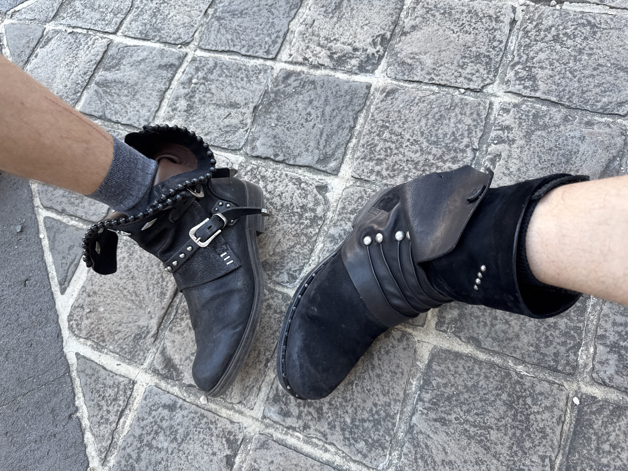 A low-angle shot captures two feet wearing black, studded, ankle-high boots on a cobblestone pavement. The left boot has a decorative, studded edge and a silver buckle. The right boot has a combination of suede and leather, also adorned with studs. The visible legs are bare and fair-skinned, with a gray sock appearing near the top of the left boot. The setting is a gray cobblestone street. The image was taken outdoors during the day. Based on the cobblestone and overall appearance, the location is likely a historic street in Paris, France.