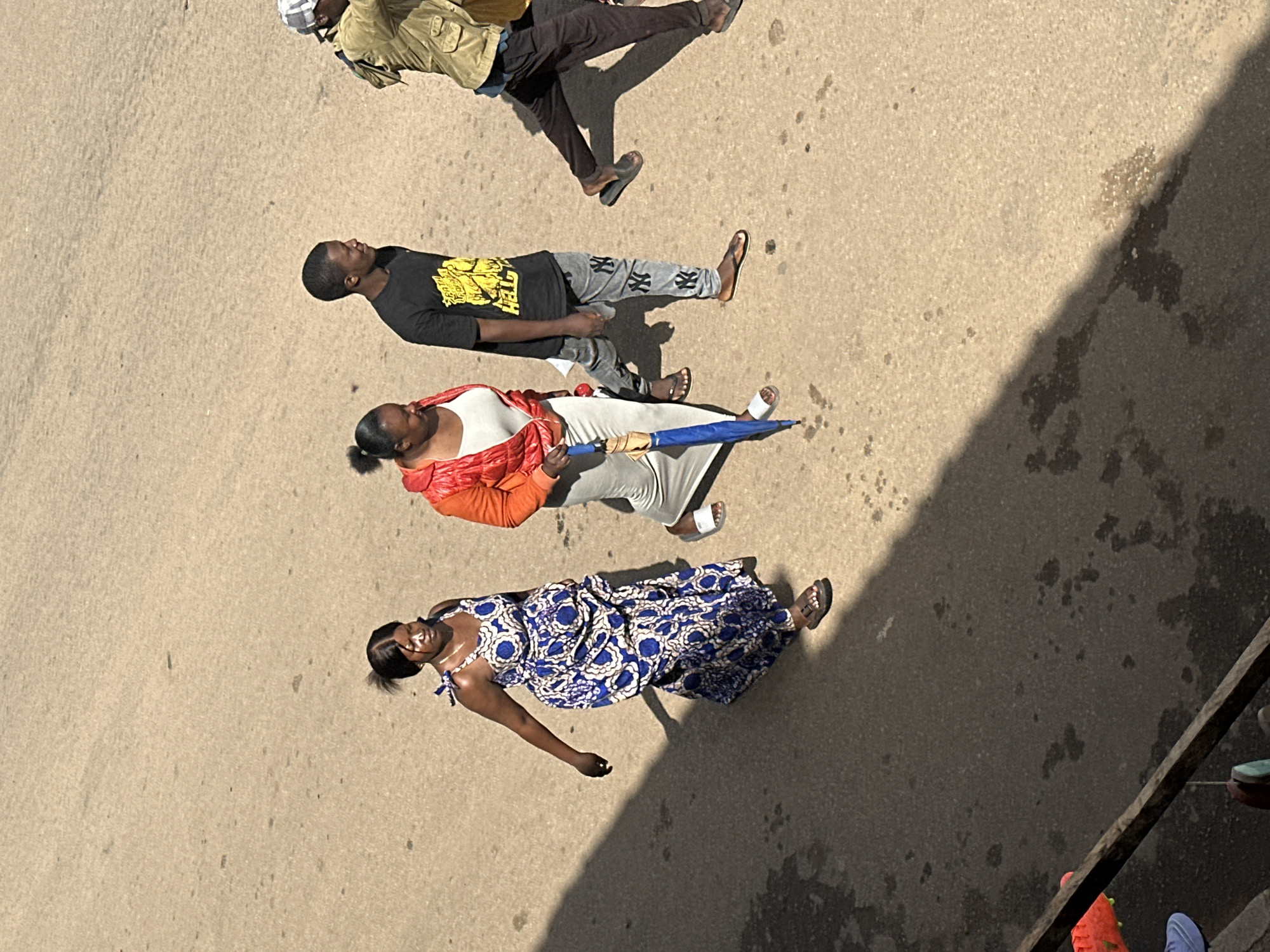 In Bukuru, Nigeria, four individuals walk across a paved surface, likely a road or pathway. The first person, a woman, is on the left, wearing a long blue and white patterned dress. She is walking with her right arm outstretched. Next to her, a woman in an orange jacket and white top is walking, holding a closed blue umbrella. To her right, a man in a black t-shirt with yellow text and gray pants with "NY" logos walks, looking to the right. Further right, another man, partially visible, is wearing a light brown jacket and dark pants, also moving to the right. The ground is a light brown, textured pavement with scattered dark spots. A shadow from the left indicates a bright light source, possibly the sun, casting a long shadow across the scene. The overall setting appears to be an outdoor urban environment.