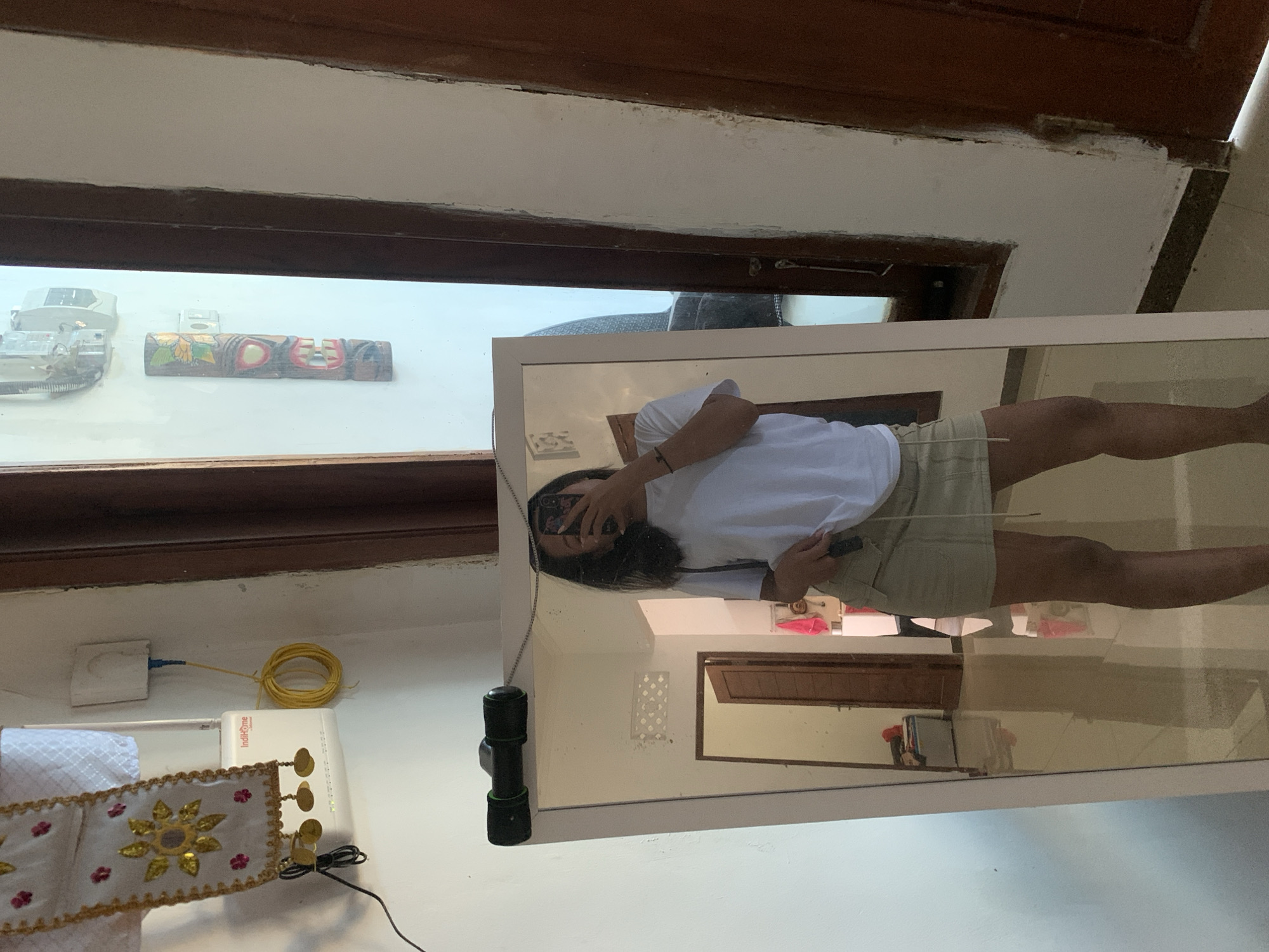 In a room in Sukawati, Indonesia, a young woman stands in front of a full-length mirror, taking a selfie with her phone. She is wearing a white t-shirt and a light brown skirt. The mirror reflects the room behind her, revealing a doorway, some furniture, and decorative items on the wall. The room appears to be residential, with tiled floors and white walls. A router is mounted on the wall near a decorative fabric hanging.