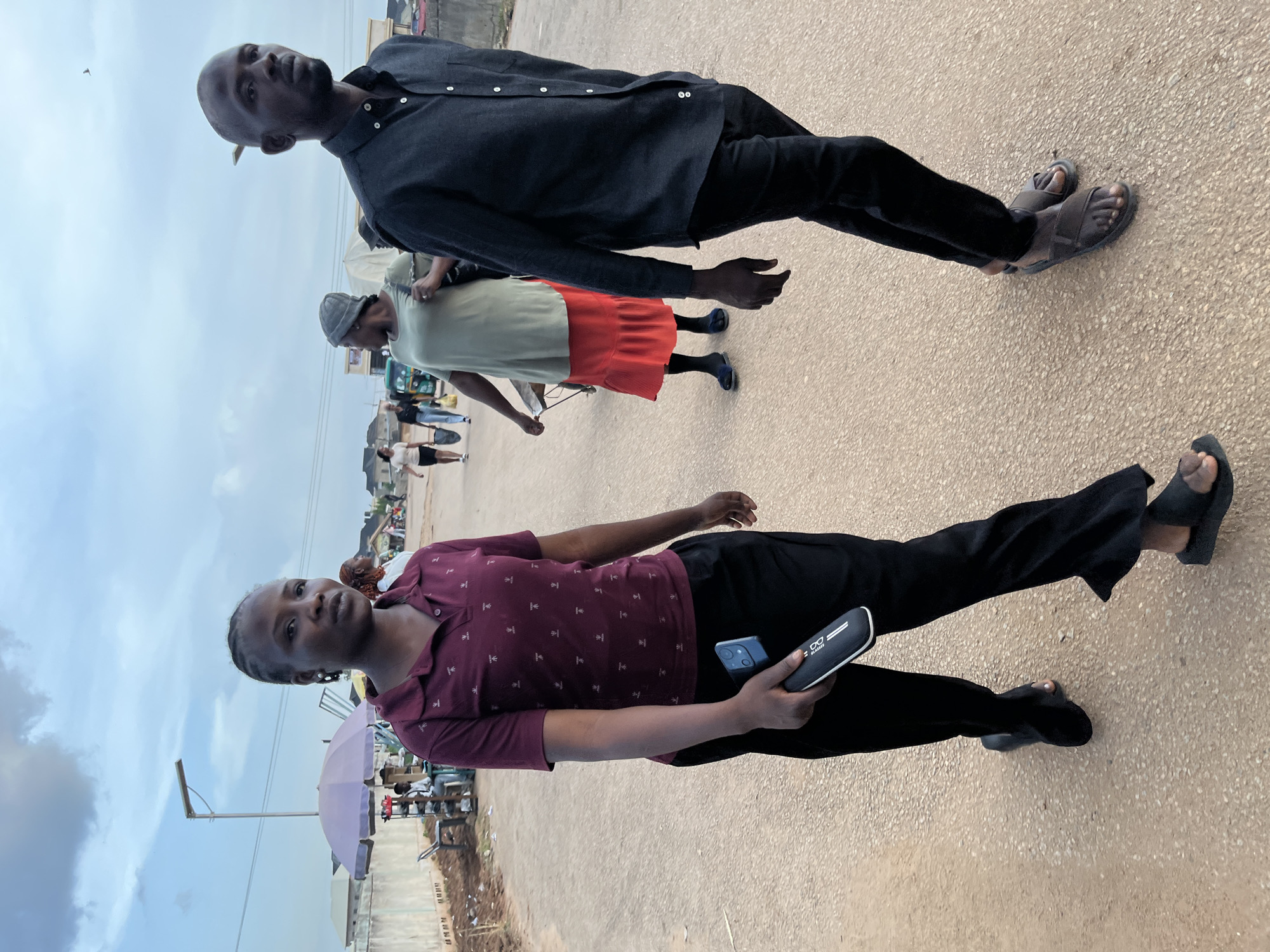The image captures a bustling street scene in Rafin Par, Nigeria, under an overcast sky. Two main figures are prominent in the foreground, both looking towards the viewer.
On the left, a woman with braided hair wears a maroon short-sleeved polo shirt with a light-colored repeating pattern, black trousers, and black slide sandals. She holds a light blue smartphone in her left hand and a black eyeglasses case, featuring a white eyeglasses icon and the text "GLASSICS," in her right hand. Her expression is neutral.
On the right, a man with very short hair or bald wears a dark charcoal or black long-sleeved collared shirt, black trousers, and dark grey slide sandals. He stands with a neutral expression, also looking towards the camera.
In the mid-ground, behind the man, another person is visible wearing a light-colored shirt and an orange or red skirt, along with dark sandals. Further back, several other individuals populate the scene, some appearing to be walking or standing near market stalls. One person wears a grey bucket hat. A light purple umbrella, likely providing shade for a vendor, is visible, along with utility poles and glimpses of distant buildings and a green tricycle (Keke Napep).
The ground is a light brown or tan paved surface, possibly asphalt or concrete. The overall impression is a daytime scene on a busy street or market area, characterized by everyday activity. The cloudy sky suggests an overcast day.