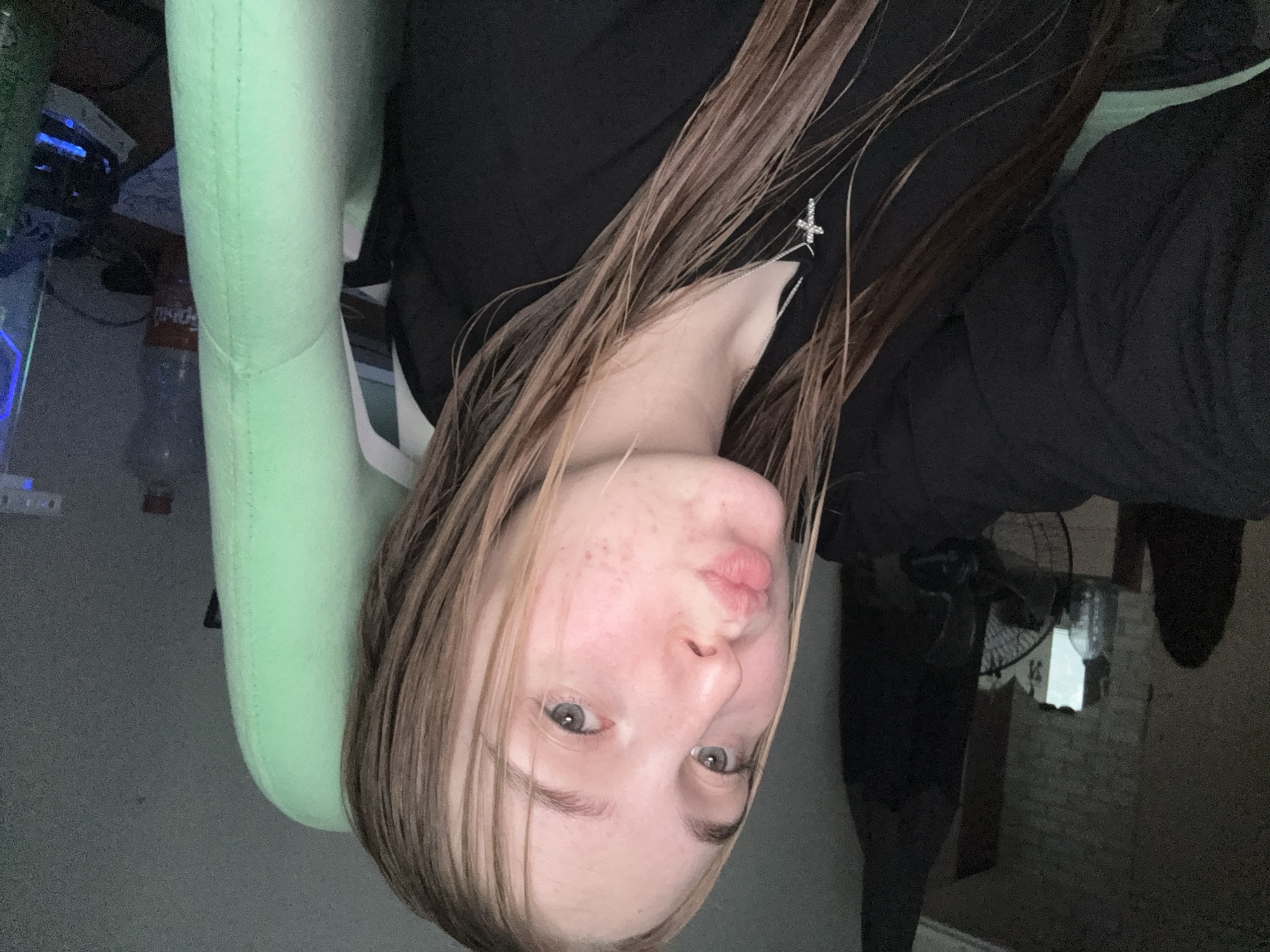 The image is oriented sideways (rotated 90 degrees clockwise), presenting a close-up selfie of a young person. She has light brown or blonde hair, which appears wet or slightly damp, falling across her face and shoulders. Her light-colored eyes are looking towards the camera, and her lips are pursed in a slight pout. Visible on her cheeks and chin are some minor skin blemishes or redness. She is wearing a black top or hoodie, and a small, silver-colored cross pendant hangs from a thin chain around her neck.
The person appears to be seated in a distinct light green (possibly mint-colored) gaming or office chair, the back and armrest of which are prominently featured. To the left side of the person (top of the image in its current orientation), the background reveals a cluttered desk or surface. Items visible include electronic devices, one emitting a blue light, and a clear plastic water bottle with an indistinguishable label.
To the right of the person (bottom of the image), the background is darker and less distinct. A partially visible fan with a white base and dark blades can be seen, along with a light source behind it that could be a window or a lamp. A section of a light-colored brick wall is also visible. The overall lighting suggests an indoor setting, likely a personal room such as a bedroom or home office, with dim light, possibly indicating evening or night. The scene is located in Perm, Russia.