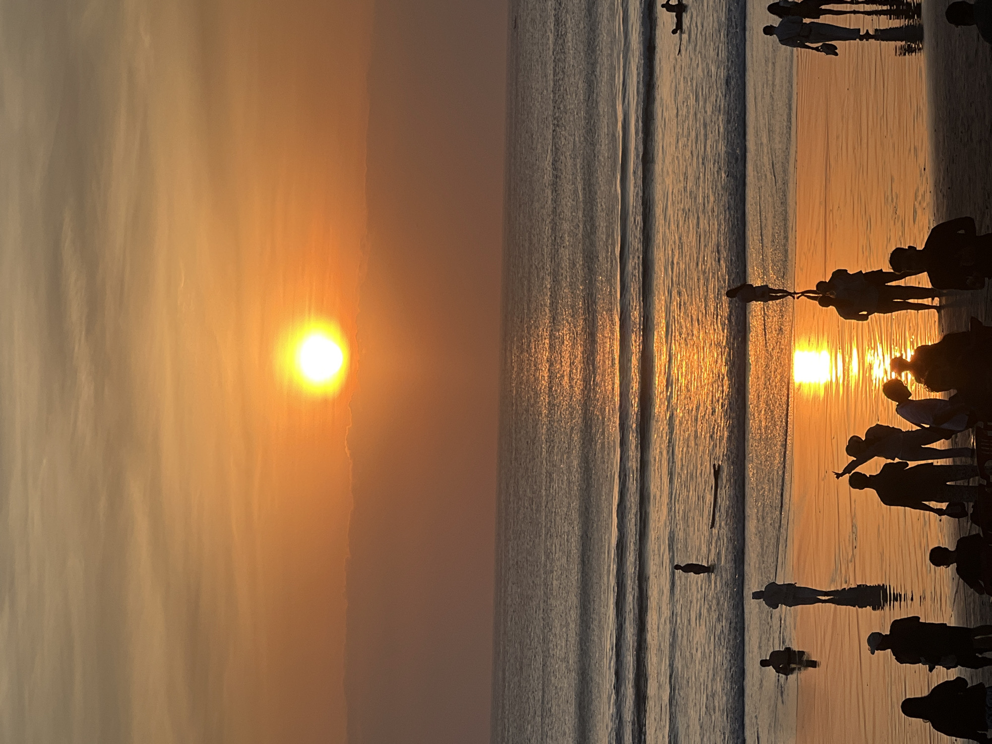 This is a vertical, wide-angle, eye-level shot of a sunset on Kuta Beach, Bali, Indonesia. The sun is a bright, diffused orb in the upper center of the frame, casting a golden-orange glow across the sky and reflecting vibrantly on the wet sand and the ocean. The water has small, foamy waves rolling towards the shore. Silhouetted figures of people are scattered across the beach and in the shallow water. In the foreground, a group of people are gathered, some standing, some seated, all in silhouette. Further back, individuals stand in the water, and some are walking along the shoreline. One person is lying on a surfboard in the ocean. The overall impression is of a peaceful, serene evening at a popular beach destination.
