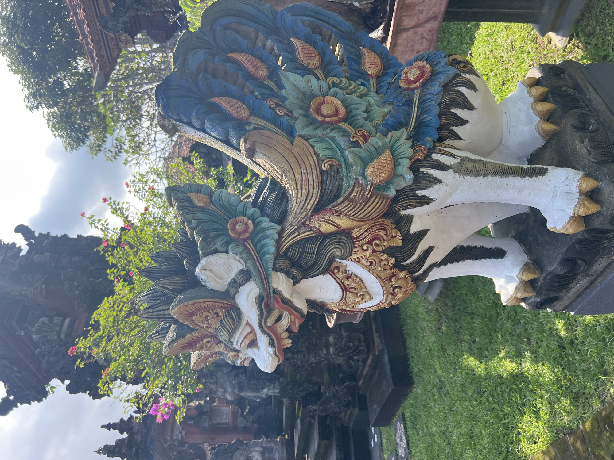 The image depicts a colorful statue of a mythical creature, likely a Barong, in what appears to be a temple or garden setting in Sukawati, Indonesia. The Barong statue is elaborately decorated with floral and feather-like motifs in shades of blue, green, gold, and red. The creature has a fierce facial expression with prominent teeth and intricate carvings. The statue is positioned outdoors, with lush green grass and plants surrounding it. In the background, there are glimpses of traditional Balinese architecture, including a temple structure with ornate carvings. The sky is visible in the background, indicating that it is daytime. The statue is the primary subject, and the setting provides context, suggesting it is a cultural or religious site in Sukawati, Indonesia.