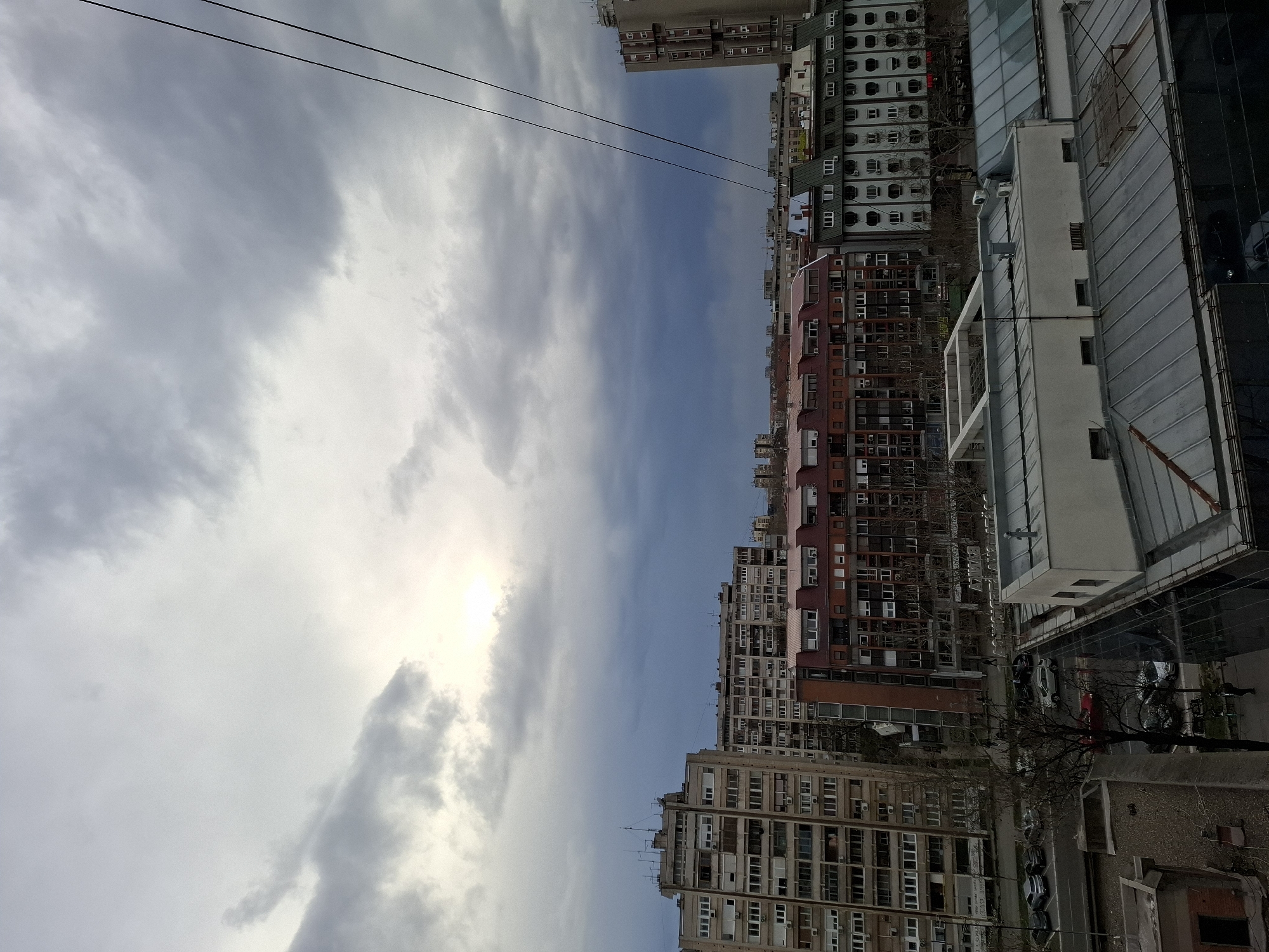 The image displays an elevated urban landscape in Novi Sad, Serbia, under a heavily clouded, grey sky. A brighter patch of light is visible through the clouds in the upper-left quadrant. Two dark utility wires extend diagonally across the upper-right portion of the frame.
In the immediate foreground, a series of rooftops are visible, including a large grey-paneled pitched roof, a flat white roof with HVAC units, and a dark, reflective glass building facade.
The midground features a multi-lane street with vehicular traffic; several cars are parked, and others are in motion. Bare deciduous trees line the street, and multiple pedestrians are visible walking on the sidewalks.
Building structures in the midground include:
* On the left, a tall, multi-story concrete residential building with numerous balconies, windows, and rooftop antennas.
* Centrally, a row of buildings consisting of a structure with a dark red tiled roof and a red-brown facade, and an adjacent building with a light-colored facade featuring distinctive rounded windows/balconies and a dark green pitched roof.
* To the far right, a portion of another tall, multi-story concrete building is visible.
The background presents a dense array of additional multi-story urban buildings, extending towards the horizon. No specific interactions between individuals or unique actions are discernible beyond general urban movement.