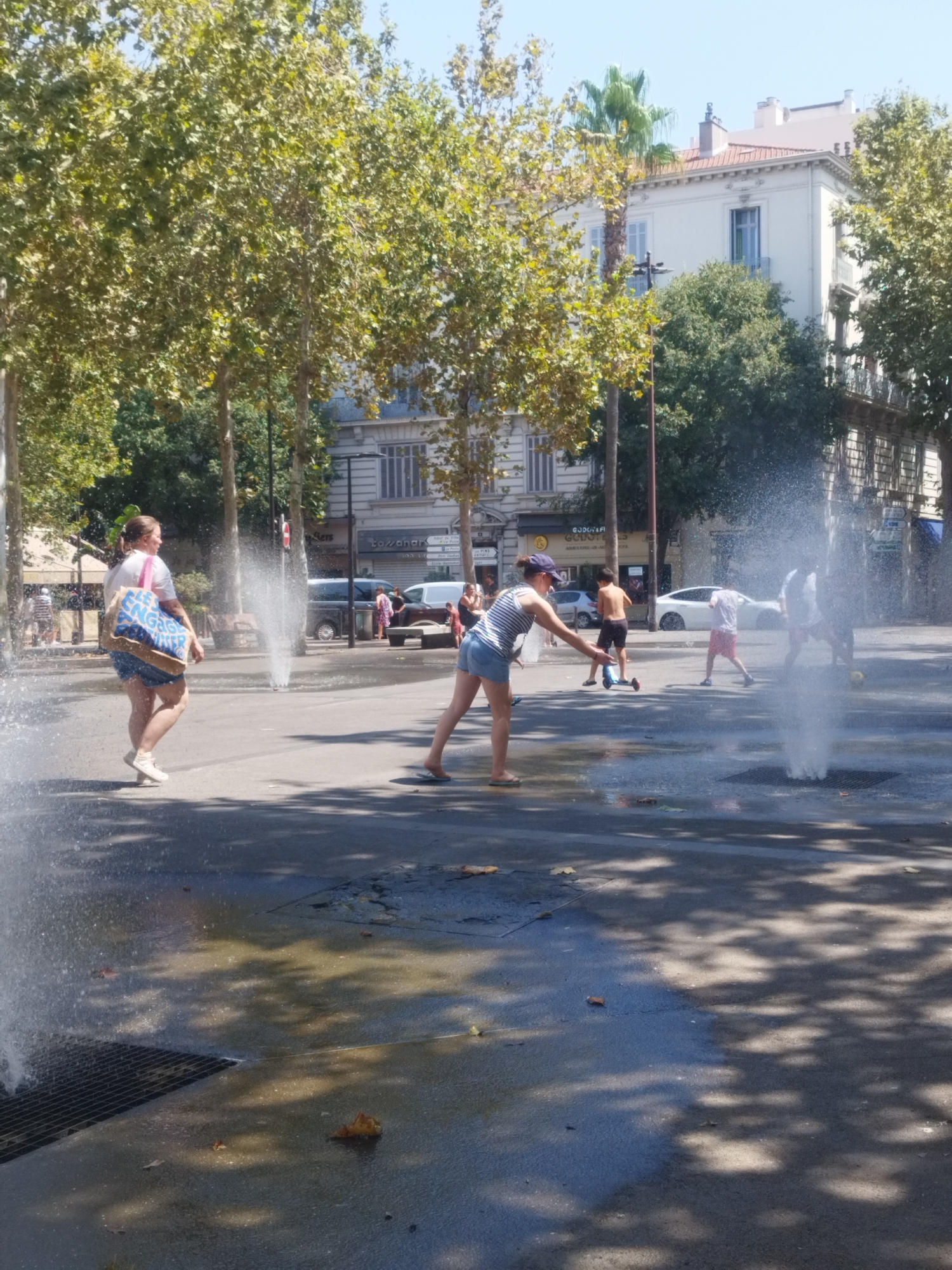 The image depicts a sunny daytime scene in a public plaza, likely in Antibes, France, with a fountain spraying water. In the foreground, a woman with her back to the camera walks away from a fountain, carrying a blue and white tote bag. She is wearing a white t-shirt and blue shorts. To her right, another woman, partially visible, is leaning forward, possibly interacting with the water. Further in the background, several individuals are scattered across the plaza. A young boy is seen riding a scooter, and another boy is wearing shorts and a t-shirt. A white car is parked behind some of these individuals. The plaza has trees lining the perimeter, and buildings with traditional architecture are visible in the background. The ground is wet from the fountain, reflecting the sunlight and casting shadows from the trees.