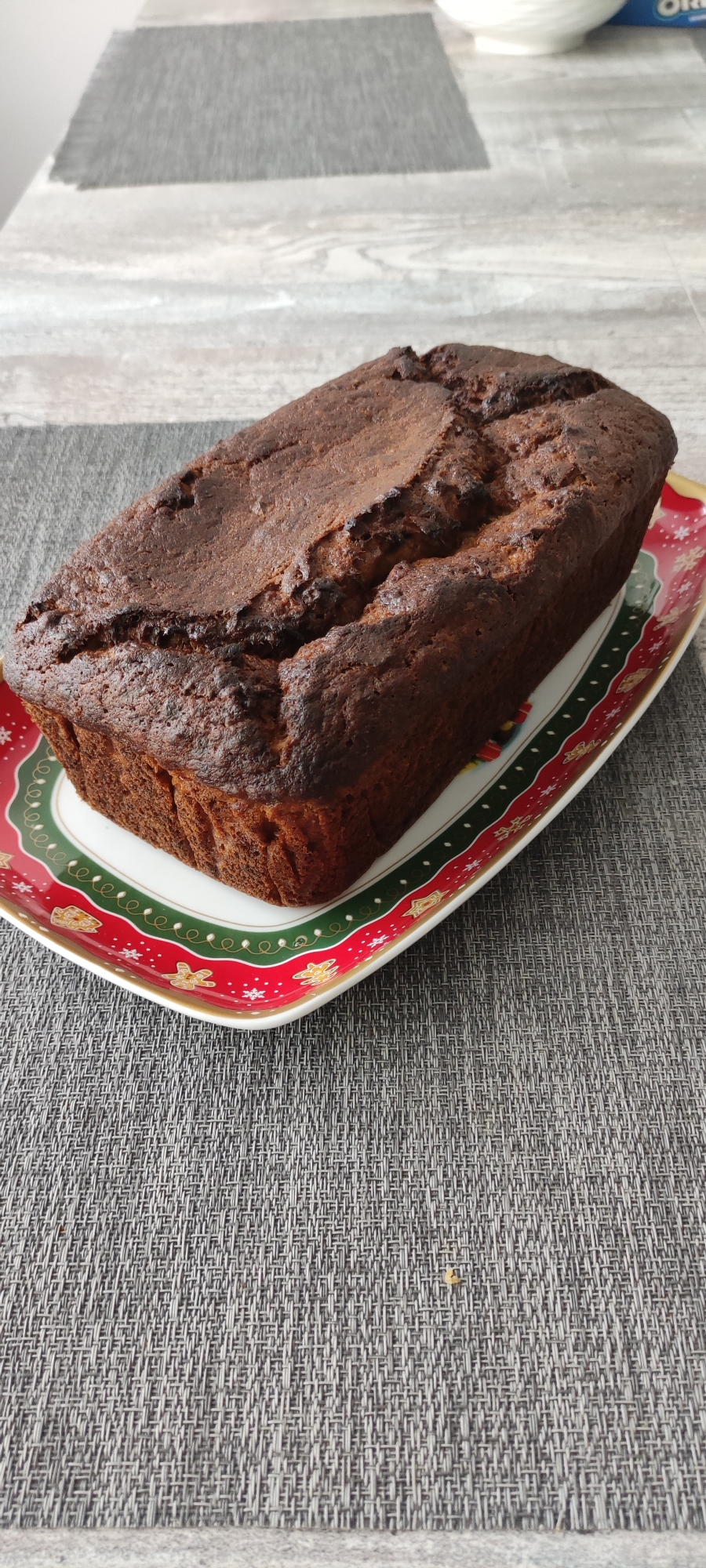 A loaf of dark brown cake is centered on a rectangular plate with a festive, red, green, and white border adorned with holiday motifs. The cake exhibits a slightly uneven, cracked top, suggesting it has recently been baked. The plate is placed on a textured gray placemat. The background consists of a gray and white patterned surface, possibly a table, with a hint of a white object and blue packaging in the upper right corner. There are no people or animals present in the image. The scene appears to be indoors, in a domestic setting.