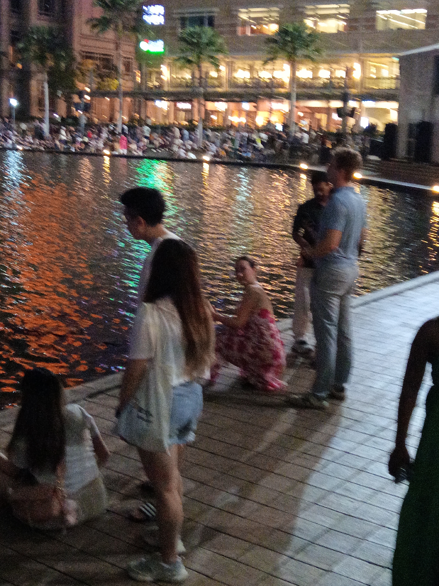 This image depicts an evening scene at a public area with a reflective body of water, likely a water feature or a canal, in front of a multi-story building. The building's windows are illuminated, suggesting it is a commercial or residential space, and there are palm trees visible in the foreground. A crowd of people is gathered along the edge of the water, some sitting and others standing, facing the water. The water's surface reflects the lights from the building and appears to have colored lights within it, creating a dynamic visual effect.
The foreground of the image shows a wooden boardwalk or pathway with several individuals. To the left, a person with long dark hair is seated, wearing a light-colored top and shorts. Further to the right, a person in a white top and denim shorts stands, with their back partially turned. Two men are standing together in the center of the frame; one is wearing a light blue t-shirt and grey pants, while the other is wearing a darker shirt and lighter pants. A woman in a patterned dress is crouching near them. The overall atmosphere is that of a public gathering or event, possibly a promenade or a place for evening relaxation.
This scene is identifiable as **Jalan Raja Laut in Kuala Lumpur, Malaysia**, specifically in the vicinity of the area with the water feature and the illuminated buildings, a common urban landscape within the city known for its vibrant nightlife and public spaces.