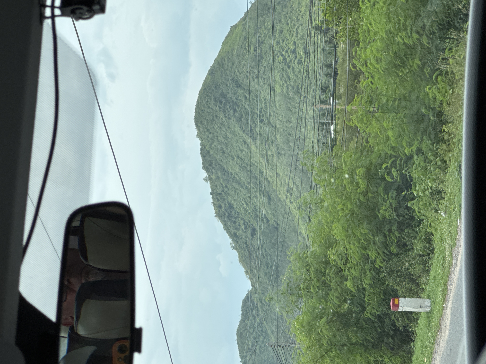 The image, taken from inside a vehicle, shows a view of rolling green hills covered in dense vegetation under a cloudy sky. In the foreground, a paved road runs along the left side of the frame, bordered by a grassy verge. A white concrete marker with red and yellow bands stands on the verge. A dense thicket of green foliage lines the road and extends towards the hills. Several horizontal power lines are visible crisscrossing in front of the hills. A rearview mirror is mounted on the windshield, reflecting a person's face and the car's interior. The overall scene suggests a rural or semi-rural location, likely outside a populated area, consistent with the countryside surrounding Ninh Ích, Vietnam.