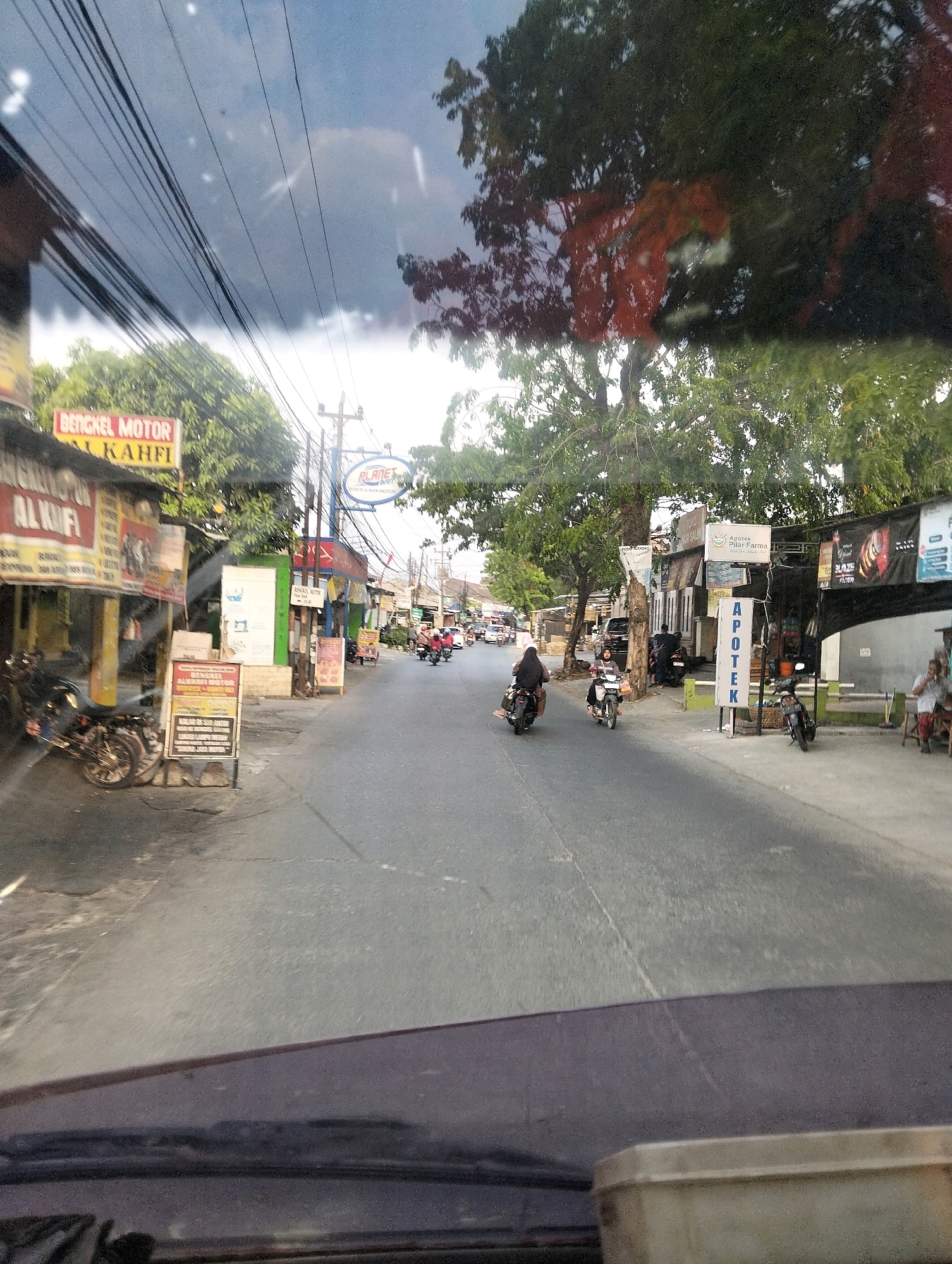 The image captures a street scene in Semarang, Indonesia, viewed from inside a vehicle. The street is lined with shops and buildings, with trees providing shade. Several motorcycles are visible, some parked and others in motion, indicating traffic. A few people are present, mostly on motorcycles. One person is seated outside a shop. The sky above is partly cloudy. Signs in Indonesian can be seen on the buildings, including one for "BENGKEL MOTOR AL KAHFI" (Al Kahfi Motorcycle Workshop) and "APOTEK PILAR FARMA" (Pilar Farma Pharmacy). The overall impression is that of a typical bustling street in a Southeast Asian city.