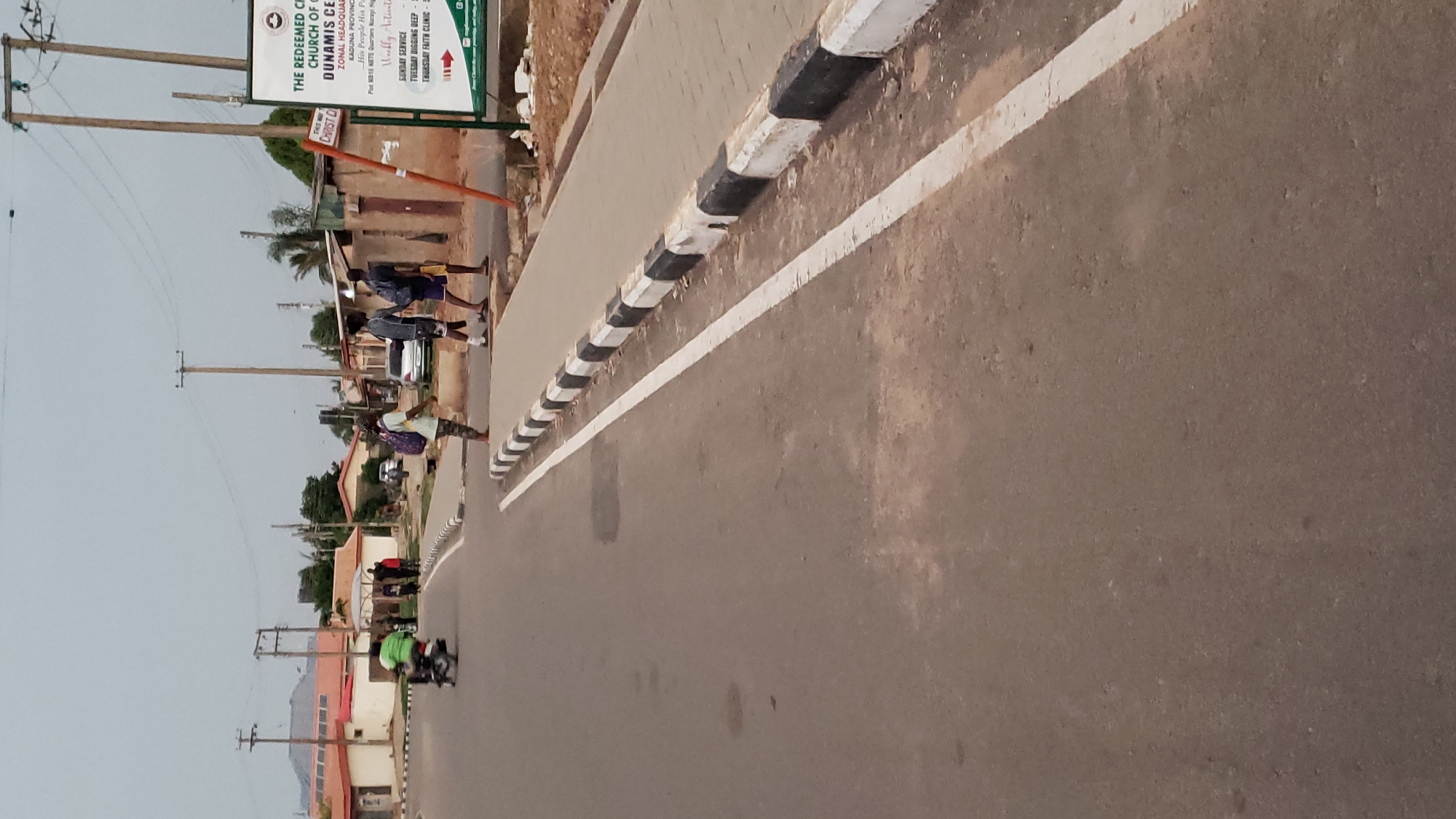 Here is a description of the image:
The image shows a street scene in Narayi High Cost, Kaduna, Nigeria. A paved road with a white line and black and white striped curb runs through the frame. People are walking along the roadside, and a motorcycle drives on the road. A sign for "The Redeemed Christian Church of God, Dunamis Centre Zonal Headquarters" is visible. Power poles are situated along the side of the road.