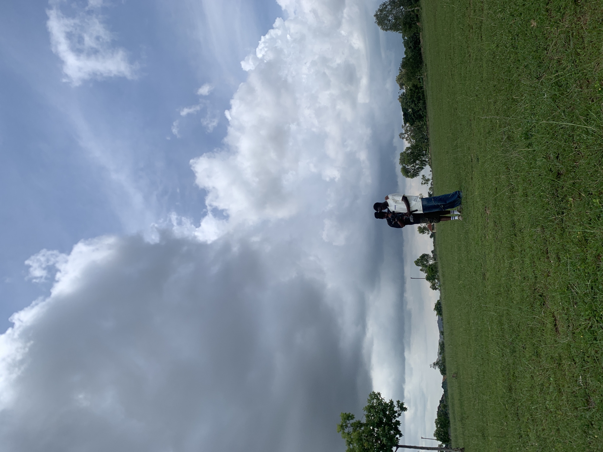 In a wide shot taken from a low angle, two young men stand in the middle of a vast, green, grassy field under a dramatic sky. The sky is a mix of dark, heavy clouds and bright white, fluffy clouds, suggesting a day with changing weather. The field stretches out in front of them, with a few trees and distant hills visible on the horizon. The men are facing each other, with one man's arm around the other's shoulder, in what appears to be a friendly or intimate pose. The man on the left is wearing a dark t-shirt and shorts, while the man on the right is in a white t-shirt and jeans. The overall impression is one of natural beauty and a moment of connection between two individuals in an open, outdoor setting. The image could be located in a park or undeveloped area within or on the outskirts of Jos, Nigeria, given the lush greenery and open landscape.