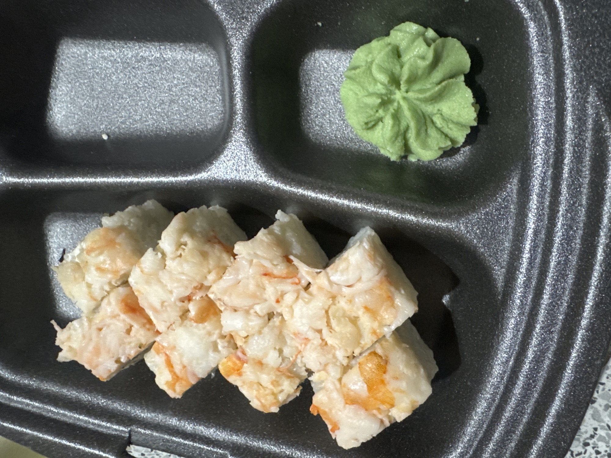A close-up overhead shot shows a black compartmentalized takeout container holding sushi and a dollop of wasabi. The container has multiple sections, with the sushi arranged in one section. The sushi pieces are cut into bite-sized portions and appear to be made with shredded or chopped seafood, possibly crab, mixed with white rice. Orange flecks, consistent with seafood, are visible throughout the sushi. In a separate compartment, a mound of bright green wasabi is neatly presented. The lighting suggests an indoor setting, possibly a restaurant or home. No people are visible in the image. The location of Zaokskiy, Russia, is not discernible from the visual information provided.
