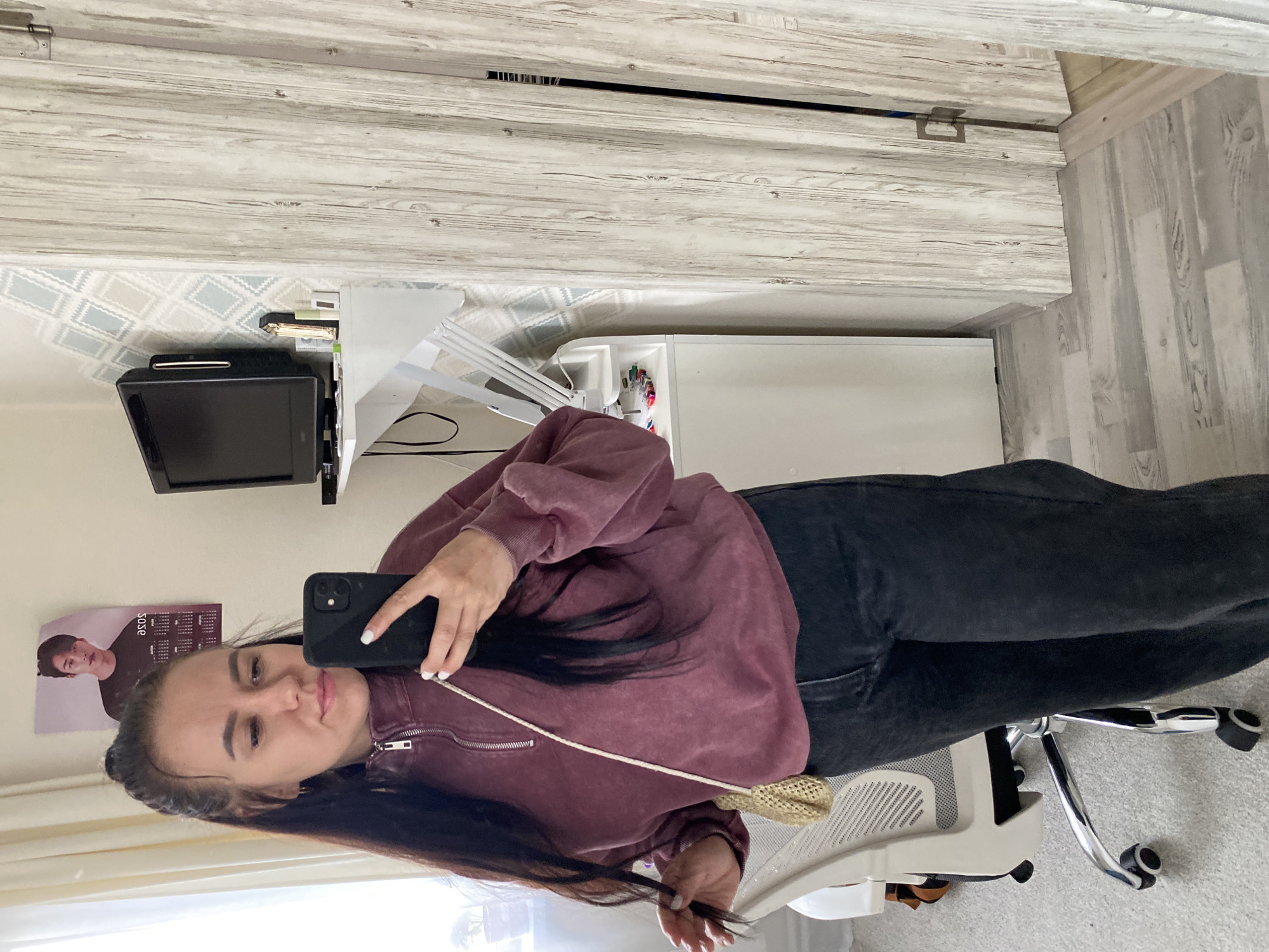 The image captures a young woman taking a selfie in an indoor setting, likely a home office or bedroom in Chaikovsky, Russia. She is seated in a light-colored office chair, holding a black smartphone in her right hand to photograph her reflection. She is wearing a maroon or burgundy quarter-zip sweatshirt and dark jeans. Her long, dark hair is styled with some strands pulled back from her face, and her fingernails are painted white. Her expression is neutral, looking towards the camera.
The background shows a white desk with a small black monitor or television on the left side. Various desk items, including what appears to be a gold stapler and other stationery, are visible on the desk surface. The wall above the desk features a light-colored wallpaper with a subtle geometric diamond pattern in shades of blue and gray. Above this, and extending to the right, are light-colored wooden panels with a distressed finish, possibly part of a wall or closet door. To the far left, on a lighter wall, a poster is visible, depicting a young man and partially showing the text "JOSE" along with a calendar grid. The floor is covered with a light-colored carpet. The overall lighting suggests it is daytime.