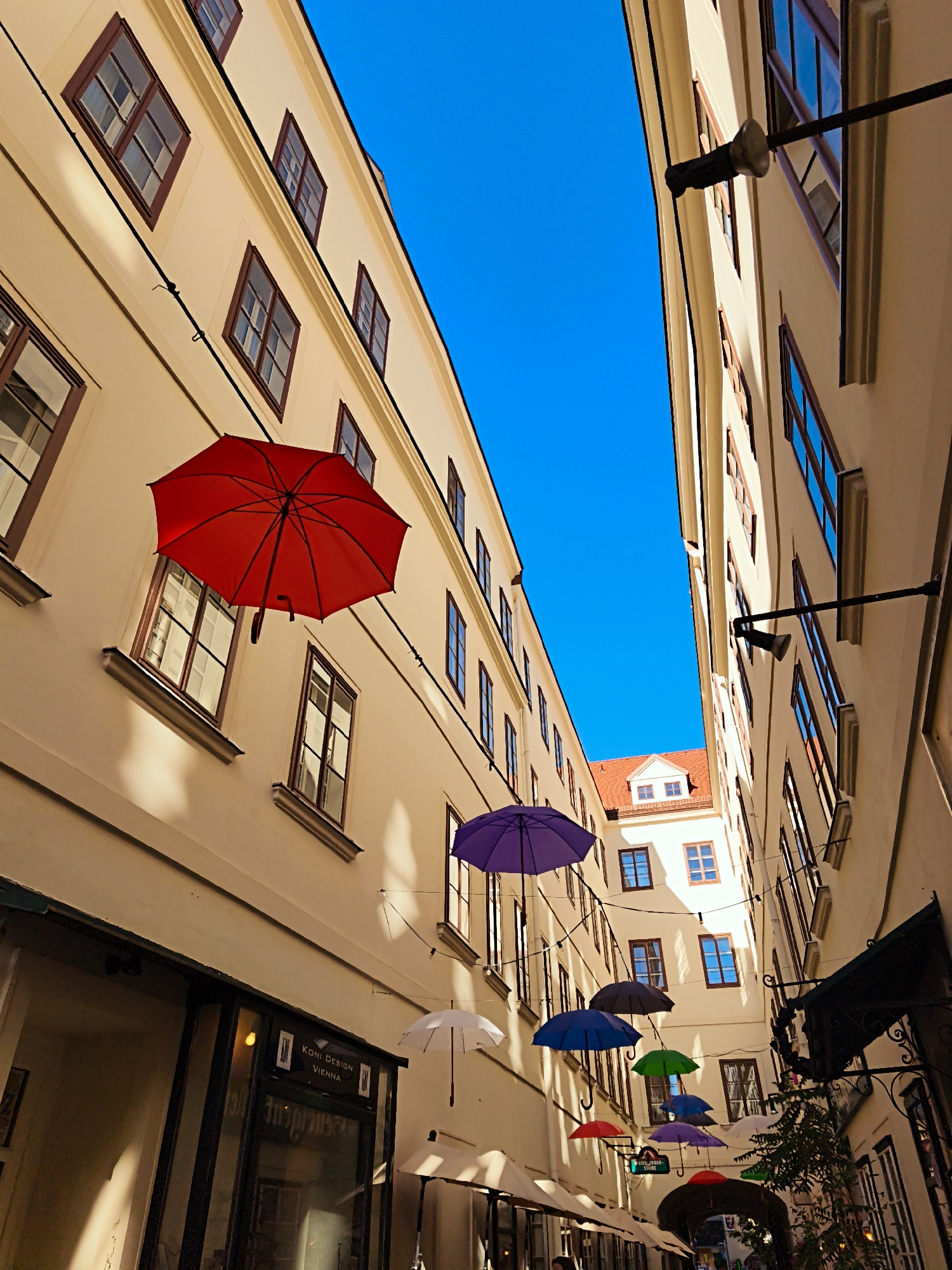 This is a low-angle, wide shot capturing a street in Vienna, Austria, lined with buildings and decorated with numerous colorful umbrellas suspended from wires. The buildings on either side of the narrow street are tall and pale yellow, with numerous dark-framed windows. A bright blue sky dominates the upper portion of the frame.
Dominating the scene are several open umbrellas in various colors: a large red umbrella is prominent on the left, a purple umbrella is slightly above and to the right of the red one, and a series of smaller umbrellas in blue, green, and purple are visible further down the street. White umbrellas are also present, one hanging on the right and others forming an awning-like structure at street level.
One building on the left side has a storefront with the words "KONI DESIGN VIENNA" visible through the glass. Shadows from the buildings and the umbrellas are cast across the facades, indicating sunlight. The perspective makes the street appear to narrow as it recedes into the distance, leading towards an arched entryway. No people are clearly visible. The scene has a festive and decorative atmosphere.