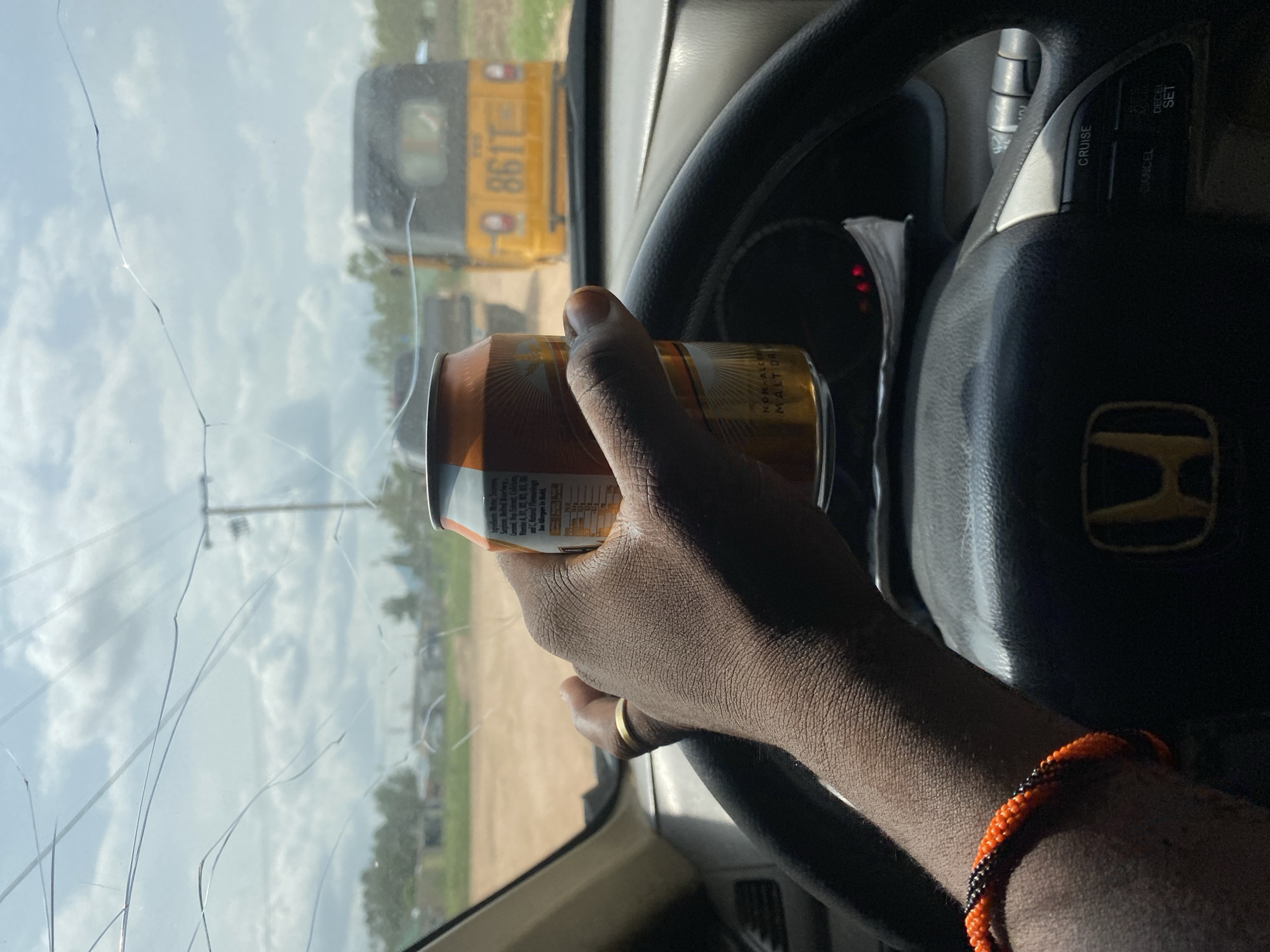 The image shows the interior of a car, with a person's hand holding a can of beverage while driving. The windshield is cracked. Outside the car, a yellow auto-rickshaw with license plate "861T" is visible, along with other vehicles and sparse vegetation. The scene appears to be in Mayo Dasa, Nigeria. The person is wearing a ring on their left hand and a beaded bracelet on their right wrist. The dashboard and steering wheel of the car are also visible.