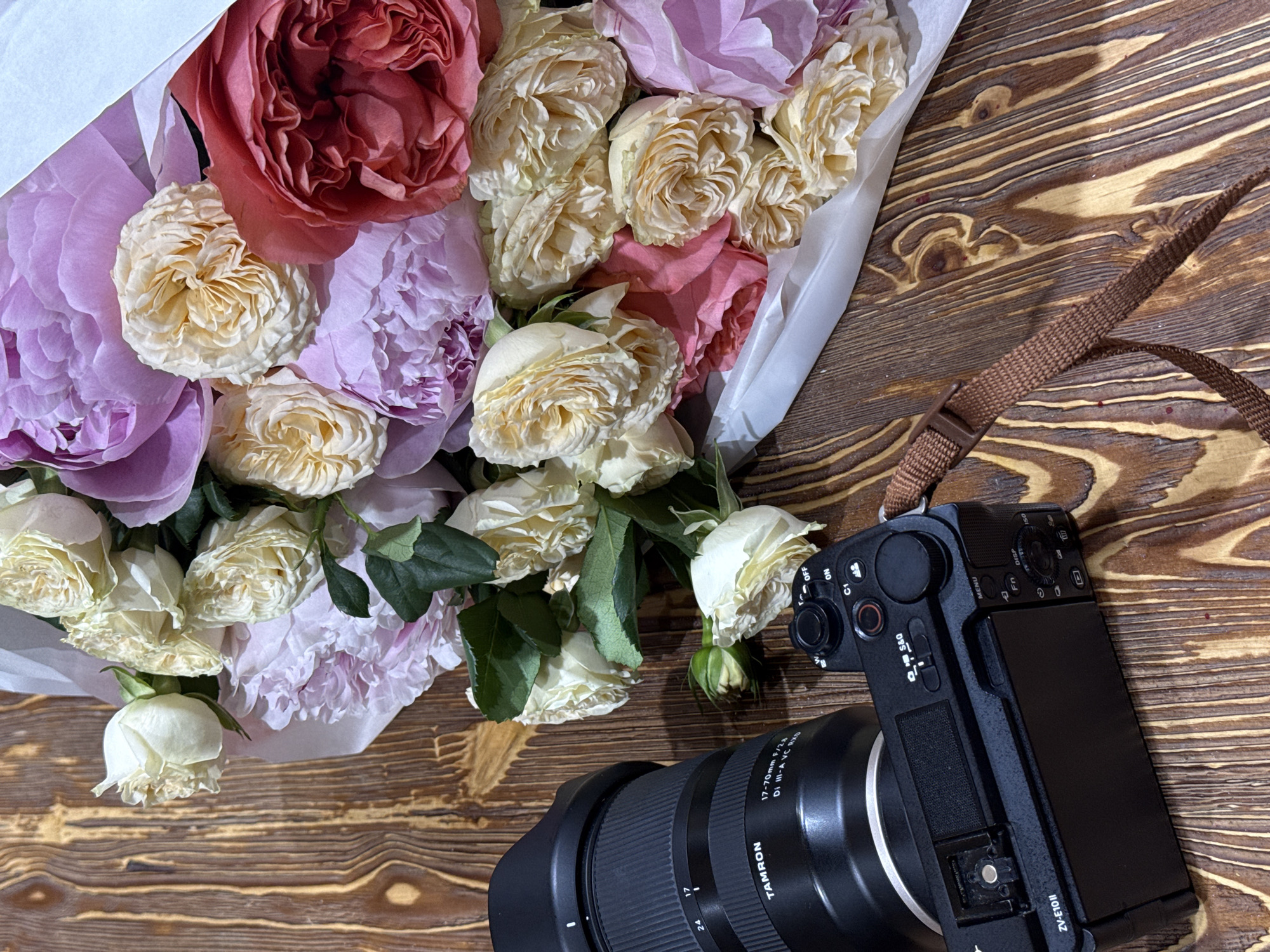 A flat lay composition displays a vibrant bouquet of flowers and a camera on a wooden surface. The bouquet, positioned on the left, features a mix of large, ruffled blooms in shades of deep red, light purple, and creamy white, resembling garden roses and peonies. The flowers are partially wrapped in white paper. Many of the petals show intricate textures and some appear slightly aged or wilted.
On the right, a black mirrorless camera with a brown fabric strap is positioned diagonally, facing towards the upper right corner. A Tamron 17-70mm F/2.8 Di III-A VC RXD lens is attached to the camera. Visible text on the camera identifies it as a "ZV-E10II" model. Other readable text on the camera and lens includes "TAMRON," "17-70mm F/2.8 Di III-A VC RXD," "S&Q," "MENU," "Fn," "DISP," "ISO," "C1," "OFF," and "ON," along with focal length markers "17" and "24" on the lens.
The background is a rustic wooden table or floor with distinct grain patterns and some small, reddish specks scattered across its surface. The lighting is soft and even, highlighting the textures of the flowers and wood.