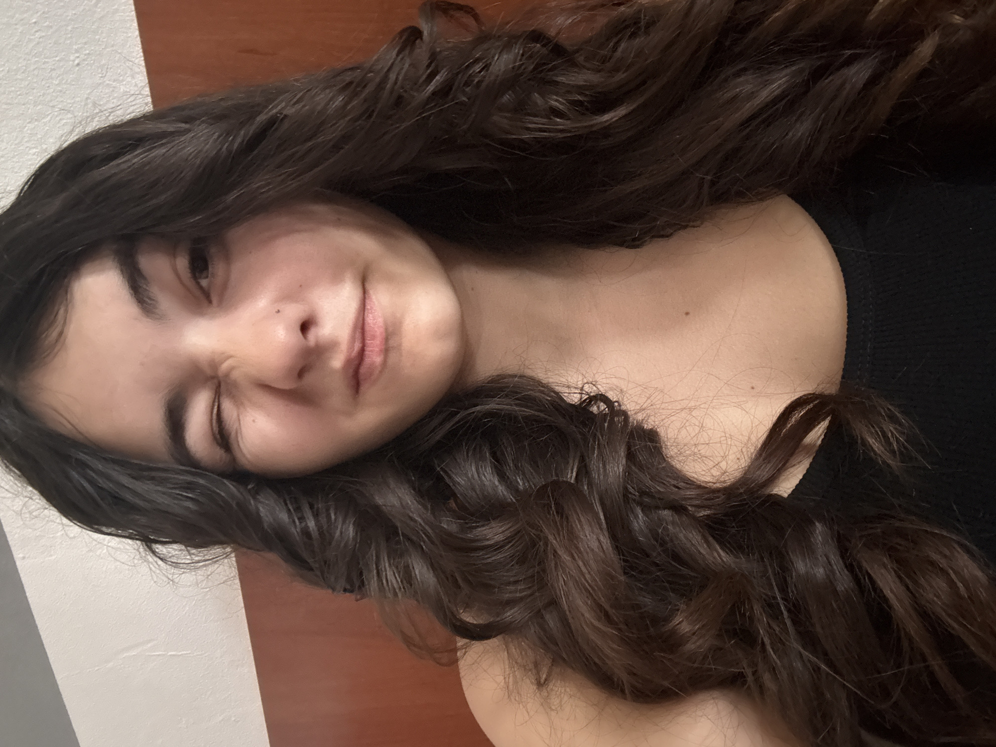 A young woman with long, dark curly hair lies on a wooden surface. She is wearing a black tank top and is looking at the camera with one eye closed. The background is blurred and out of focus. The location is unknown and cannot be determined from the image.