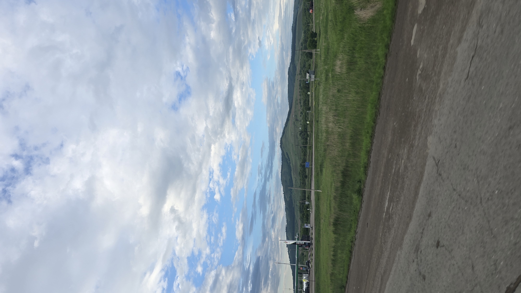 The image captures a wide-angle view of a rural or semi-rural landscape in Karabash, Russia, under a partly cloudy sky. The scene is bisected horizontally by a road and fields in the foreground and midground, leading to hills on the distant horizon.
The upper approximately two-fifths of the image consists of a sky with a mix of stratocumulus and altocumulus clouds. These clouds vary in density and shade, from bright white to light gray, with intermittent patches of clear blue sky visible, primarily in the lower part of the cloud layer.
The midground features a prominent, low-lying range of verdant hills or mountains stretching across the entire horizon. These hills are uniformly covered in green vegetation, appearing densely forested on their upper slopes. Below the hills, a horizontal band of lighter green fields and scattered shrubbery extends towards the foreground road. On the left side of the midground, a gas station is visible with a distinctive white canopy structure and green accents, sheltering several fuel pumps. Multiple vehicles are parked or positioned near the gas station, though specific details are indistinct. A paved road runs horizontally across the midground, separating the immediate fields from the more distant landscape elements. Several slender, vertical poles, likely utility or communication poles, are strategically placed along the road and within the fields. A few blue rectangular signs, possibly road signs or billboards, are discernable in the mid-distance to the right of the gas station. A faint, reddish vehicle, possibly a truck, is barely visible on the road in the far right midground.
The foreground consists of two distinct strips. The bottom left quarter shows a gray, asphalt-like surface, likely a road or a shoulder, exhibiting cracks and patches of dirt, suggesting wear. Adjacent to this paved area, to its right, is a broad strip of vibrant green, tall grass, forming a natural border along the roadside.
The lighting indicates daytime conditions, with diffused light filtering through the cloud cover. No individuals are explicitly visible or engaged in any discernible activity within the frame.
