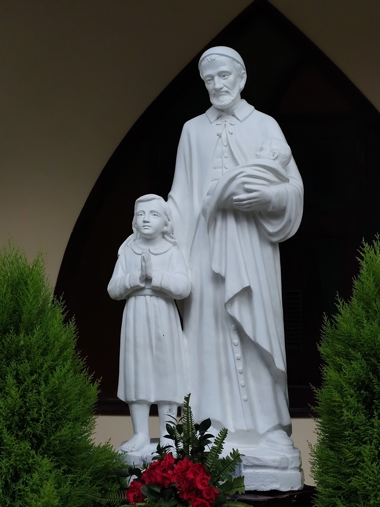 The image shows a white statue of a man, a child, and a baby in Da Lat, Vietnam. The man has a beard and is wearing a long robe, holding a baby wrapped in cloth. Next to him stands a child with hands clasped in prayer. The statue is set against a dark, arched doorway with light cream trim. Two green bushes frame the statue on either side, and red flowers are at the base.