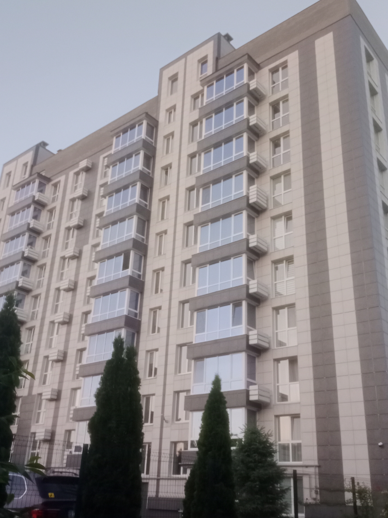 The image depicts the exterior of a modern apartment building in Horishni Plavni, Ukraine. The building is tall and features a facade with alternating sections of light-colored stone and darker gray paneling. Many windows are visible, arranged in vertical columns and in projecting bay window structures. The sky above is overcast and a pale gray. In the foreground, there are several dark green evergreen trees, with a portion of a dark-colored car visible on the left. A fence runs along the bottom of the image.