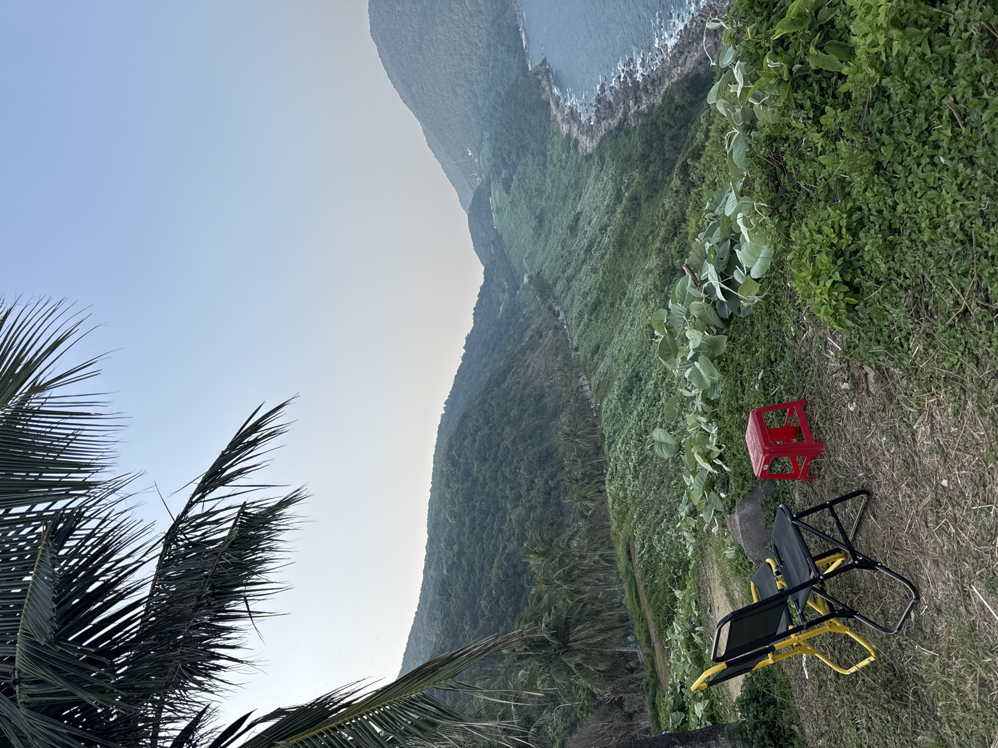 The photograph depicts a landscape with a coastline, vegetation, and seating. A red plastic stool and a folding chair with a yellow frame and black seat rest on grass. In the foreground, palm fronds partially obstruct the view. The background consists of a mountainous terrain covered with dense green vegetation that extends down to the coastline. The sky is clear and pale blue. The location is likely near a coastal area in Đà Nẵng, Vietnam, given the tropical vegetation and topography. There is no evidence of human activity in the shot other than the presence of the chairs.